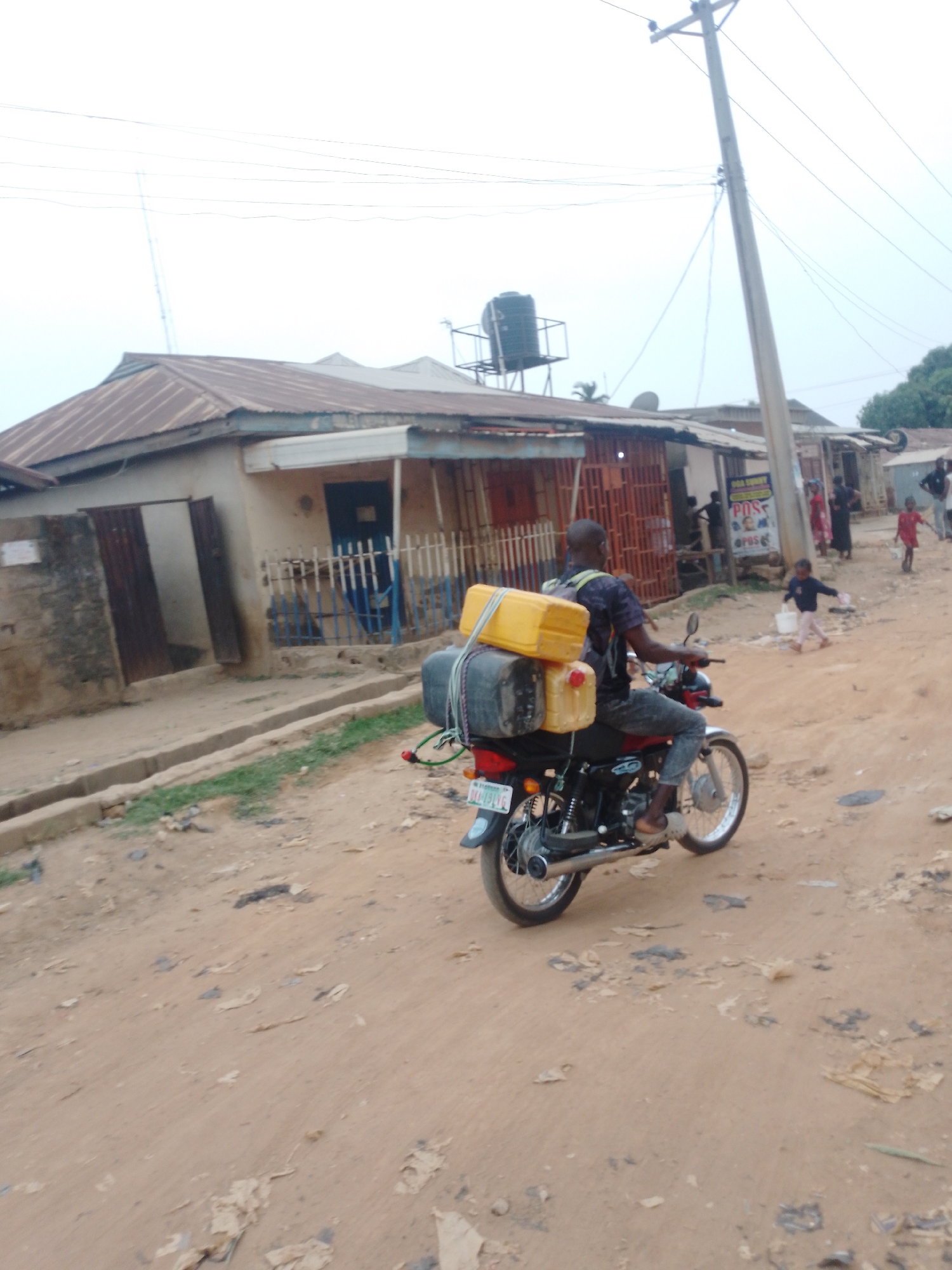 The image shows a man on a motorcycle with two yellow jerrycans and luggage strapped to the back, on a dirt road in Sabon Tasha, Nigeria. The background includes low-rise buildings with rusted metal roofs and deteriorating walls, utility poles, and a water tank on a platform. Several people, including children, are walking near a building with a sign that reads "POS". The road is unpaved and littered with debris.