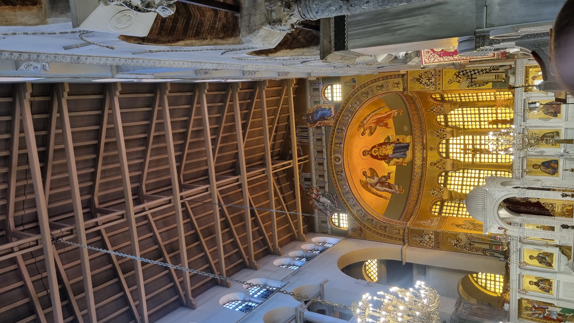 The image captures the interior of the Hagia Sophia in Thessaloniki, Greece, looking towards the altar and apse. The wooden, beamed ceiling is visible at the top, with chains hanging down. The apse features a large mosaic of the Virgin Mary holding the infant Jesus, flanked by angels. Below the mosaic are rows of arched windows illuminated from behind, casting a warm glow. The walls are adorned with further mosaics and religious icons. An iconostasis, a wall of icons, stands in front of the altar area, with a white, dome-like structure at its center. Ornate chandeliers hang from the ceiling. The overall impression is one of solemnity and grandeur, characteristic of a Byzantine basilica. No people are clearly visible in the image.