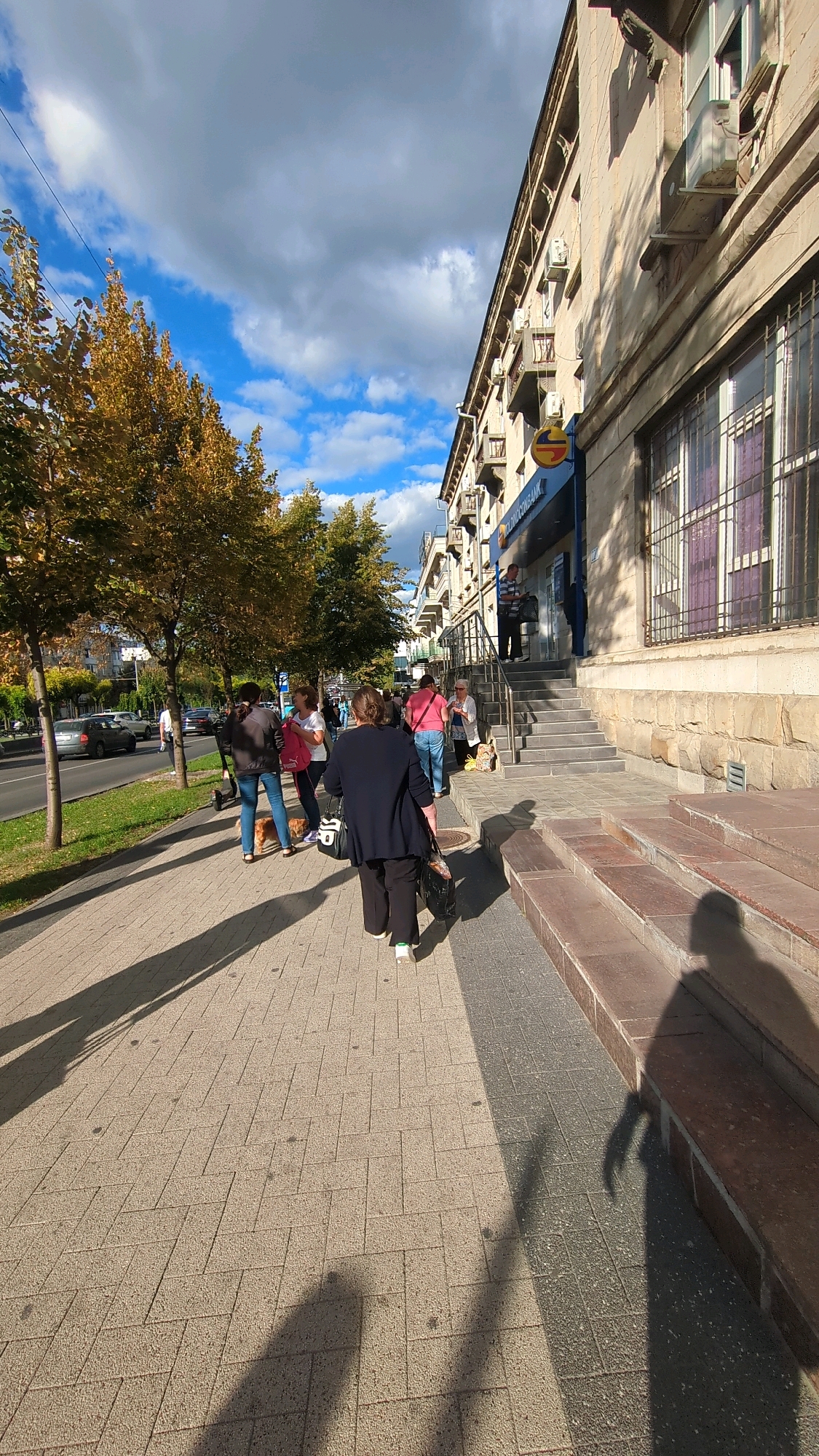 The image captures a scene on a sidewalk in Chisinau, Moldova, likely in front of a bank. The time of day appears to be afternoon, evidenced by the long shadows cast by the people and the trees. The architecture suggests a European city, with a building featuring multiple windows and air conditioning units. On the sidewalk, several individuals are present. One person, dressed in a dark top and pants, walks away from the camera. Another person, wearing a pink top and blue jeans, is standing. Further down the sidewalk, a group of people are gathered, with some appearing to be waiting in line. Trees with yellowing leaves, indicating autumn, line the left side of the frame, and a street with cars is visible beyond them. The pavement is made of interlocking concrete bricks.
