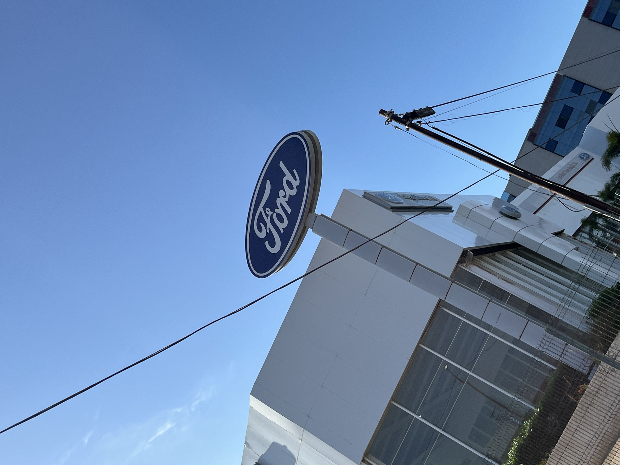 A low-angle outdoor photograph captures a prominent Ford dealership building and its sign against a clear blue sky. The central subject is a large, oval-shaped sign featuring the iconic "Ford" script logo in white against a deep blue background. This sign is mounted on a white structure that is part of the modern, multi-story dealership building.
The building itself is characterized by its contemporary architecture, utilizing white and gray paneling with large, reflective glass windows. The sunlight creates strong highlights on the white surfaces and reflections in the glass. On one section of the building, a smaller sign is visible with text that appears to read "Latin America" below a circular logo.
In the upper right portion of the frame, a utility pole with multiple wires extends diagonally across the sky and towards the building. Several wires are stretched across the clear sky. Part of a chain-link fence and some green foliage are visible near the base of the building on the right side. The bright, cloudless sky indicates a sunny day, likely morning or early afternoon. The image depicts a commercial establishment, specifically a Ford dealership, in Porlamar, Venezuela.