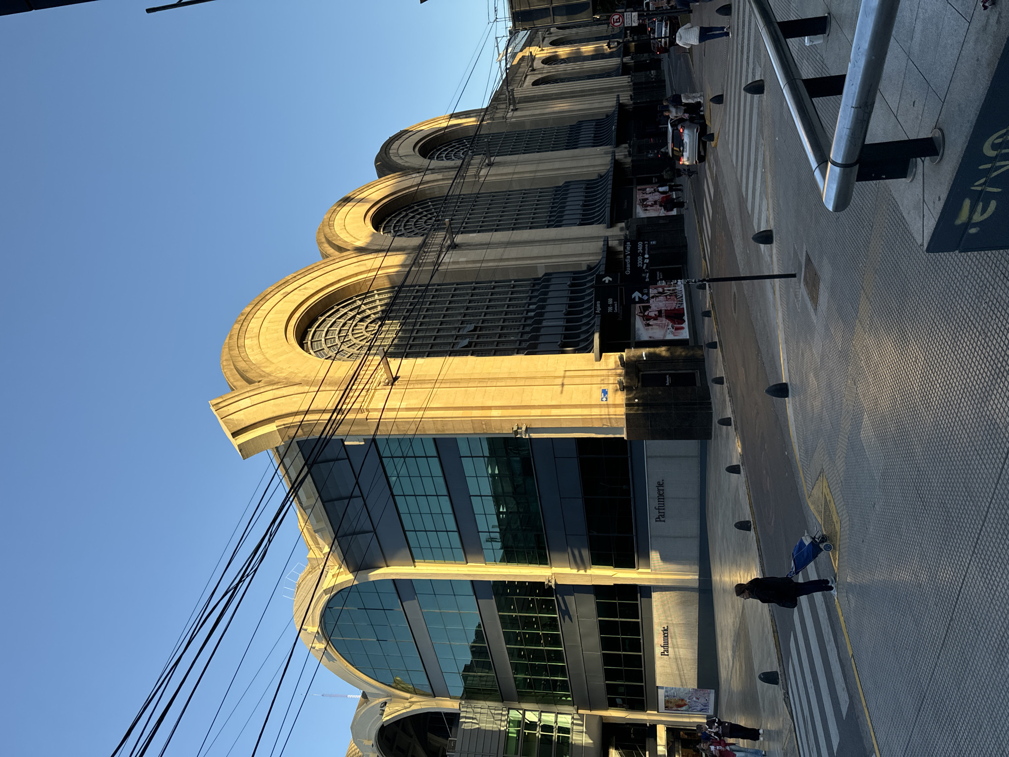 An outdoor, eye-level street view captures a grand building facade and a bustling sidewalk scene, likely in Buenos Aires, Argentina, under a clear blue sky. The building features a distinctive design with several large, prominent arched windows on its upper levels, and modern glass-fronted sections on the lower floors. The architecture, illuminated by warm, golden sunlight from the right, suggests either late afternoon or early morning, casting long shadows across the street.
Numerous black power lines crisscross in front of the building, connecting to poles unseen in the frame. On the ground level, signs for "Parfumerie." are visible on the building's exterior. The street is paved, featuring a zebra crosswalk where a pedestrian in a dark coat walks pushing a blue shopping cart. Other pedestrians are visible further down the street, and a few cars are parked or moving slowly in the background. Street furniture includes bollards lining the pavement and a metal barrier on the far right.
Notable text includes street signs in Spanish. One sign, pointing right, reads "Guardia Vieja" with address numbers "3300 - 3400" below. Another sign, pointing left, indicates "Aguero" with numbers "700 - 800". A 'no parking' symbol is also visible near vehicles in the distance. The overall scene depicts a typical urban environment with a mix of historical architecture and modern elements under bright, clear weather conditions.