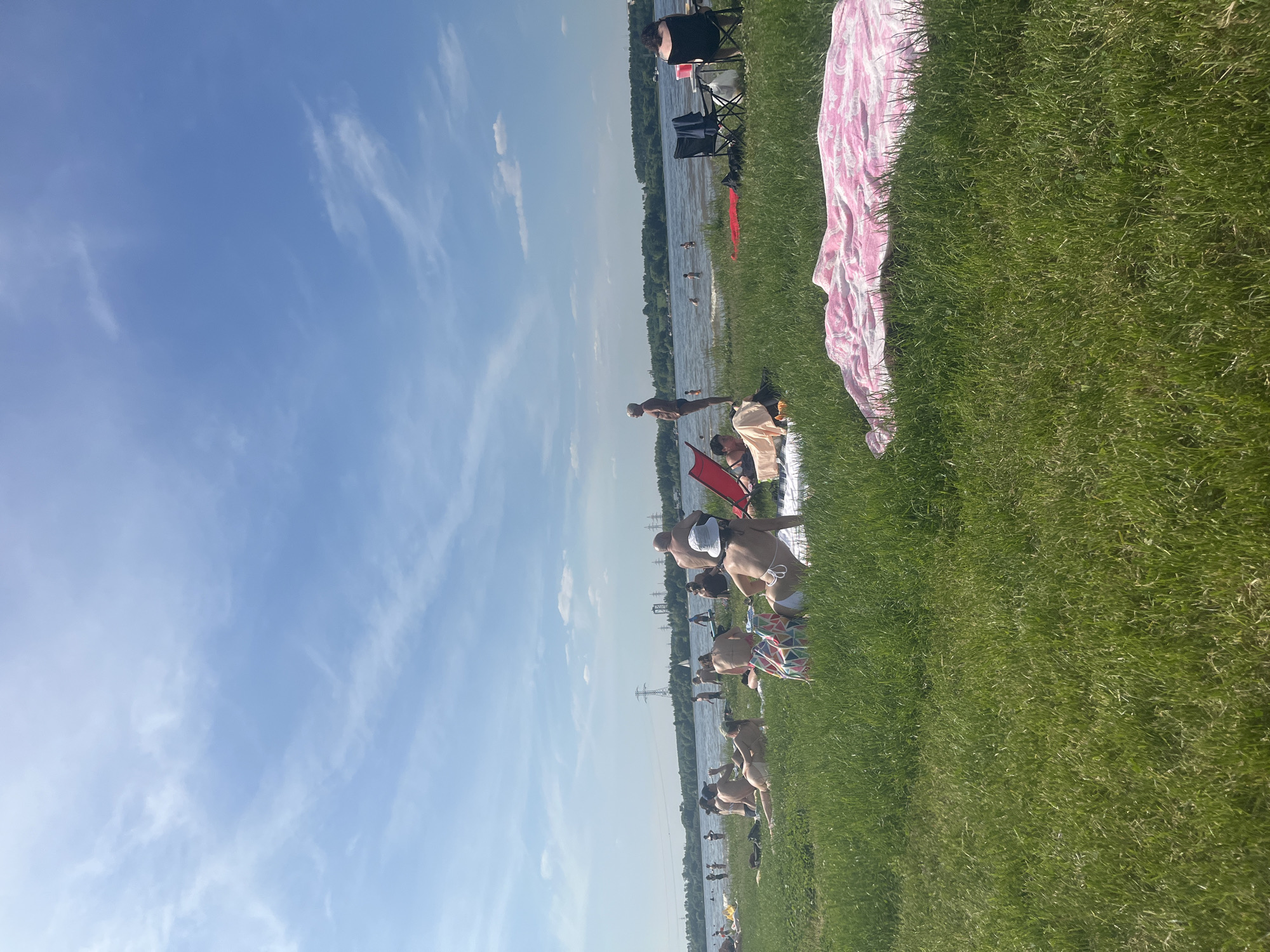 This image captures a wide shot of a public recreation area along a body of water in Kapustino, Russia, on a bright, sunny day. The scene is dominated by a clear blue sky with sparse, wispy white clouds and a large expanse of vibrant green grass in the foreground and midground.
Approximately 20-30 individuals are present on the grassy shore and in the water. Many are sunbathing or resting on the grass, some on towels. For instance, a pink and white patterned towel is visible in the lower right foreground. Several people are wading or standing in the calm, light blue water. An older male in dark swim briefs stands at the water's edge on the left side of the frame. In the mid-right, a person wearing a white bucket hat and a light-colored bikini is lying on the grass, next to a multicolored geometric pattern beach bag. Nearby, a red portable beach chair is set up, and two black portable chairs are positioned further to the right, one occupied by a person in a dark top.
Across the water in the background, a dense line of green trees marks the opposite shore. Above the treeline, multiple large metal power line towers and other indistinct industrial structures are visible. A small white sailboat is present on the water in the distant midground. Shadows on the grass suggest direct sunlight.