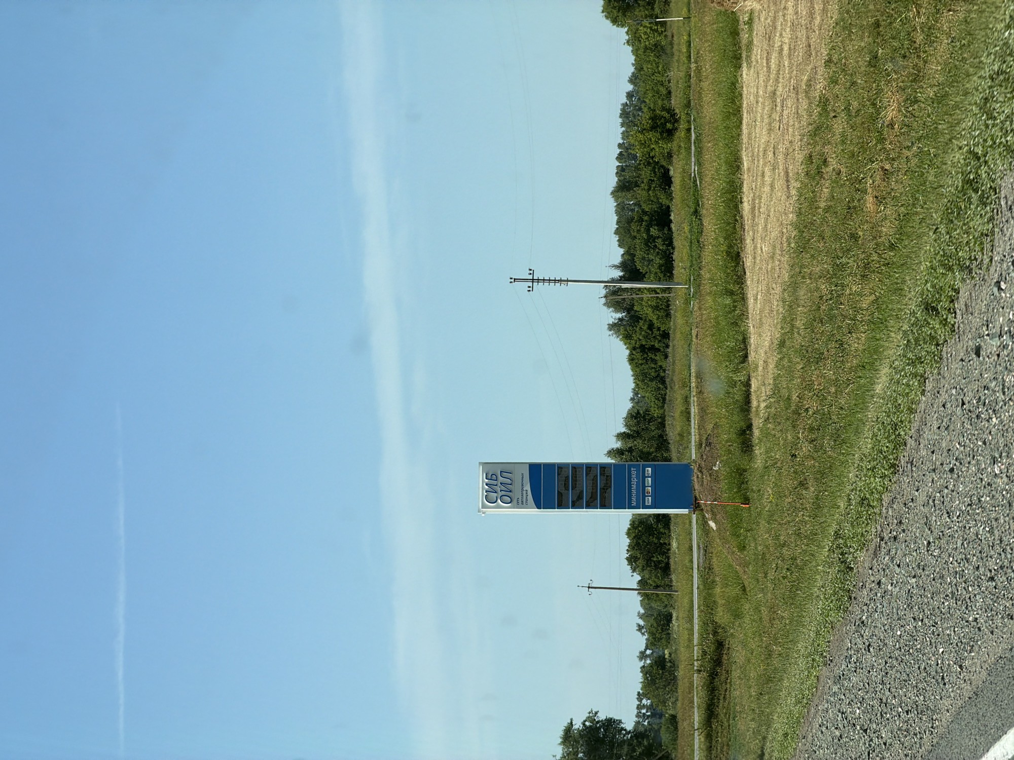 A roadside sign for a "SIB OIL" gas station is prominently displayed in a rural landscape. The sign is blue and white, with the "SIB OIL" logo at the top, followed by text that appears to indicate "service station" in Russian. Below this, there are several rectangular panels that likely display fuel prices, though the numbers are not clearly legible. At the bottom of the sign, it indicates "minimarket" and displays logos for various payment methods, including Visa and Mastercard. The sign is situated in a grassy field with a gravel road in the foreground and trees and a clear blue sky in the background. The environment suggests a sunny day in a location that could be near Bezmenovo, Russia, given the likely Cyrillic script on the sign. There are no people or animals visible in the image.