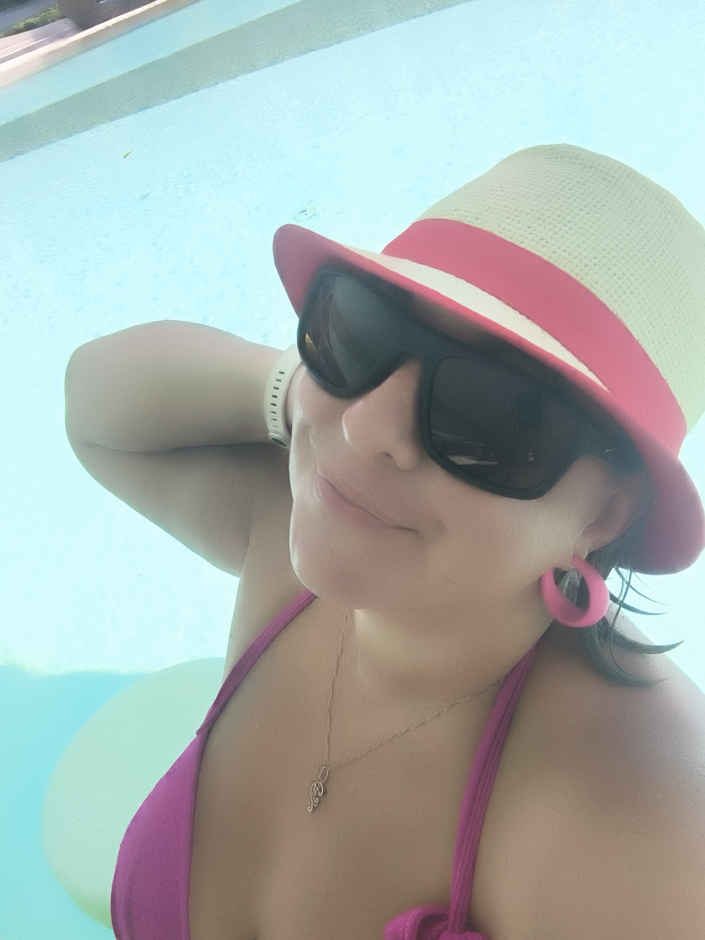 A woman is in a swimming pool, wearing a pink bikini, a straw hat with a pink band, large dark sunglasses, and pink hoop earrings. She is also wearing a white fitness tracker on her left wrist and a delicate silver necklace with a pendant. Her left arm is bent behind her head. The water is a clear turquoise color, and the edge of the pool and a portion of a pool float are visible. The scene appears to be a resort or hotel pool in Phú Quốc, Vietnam.