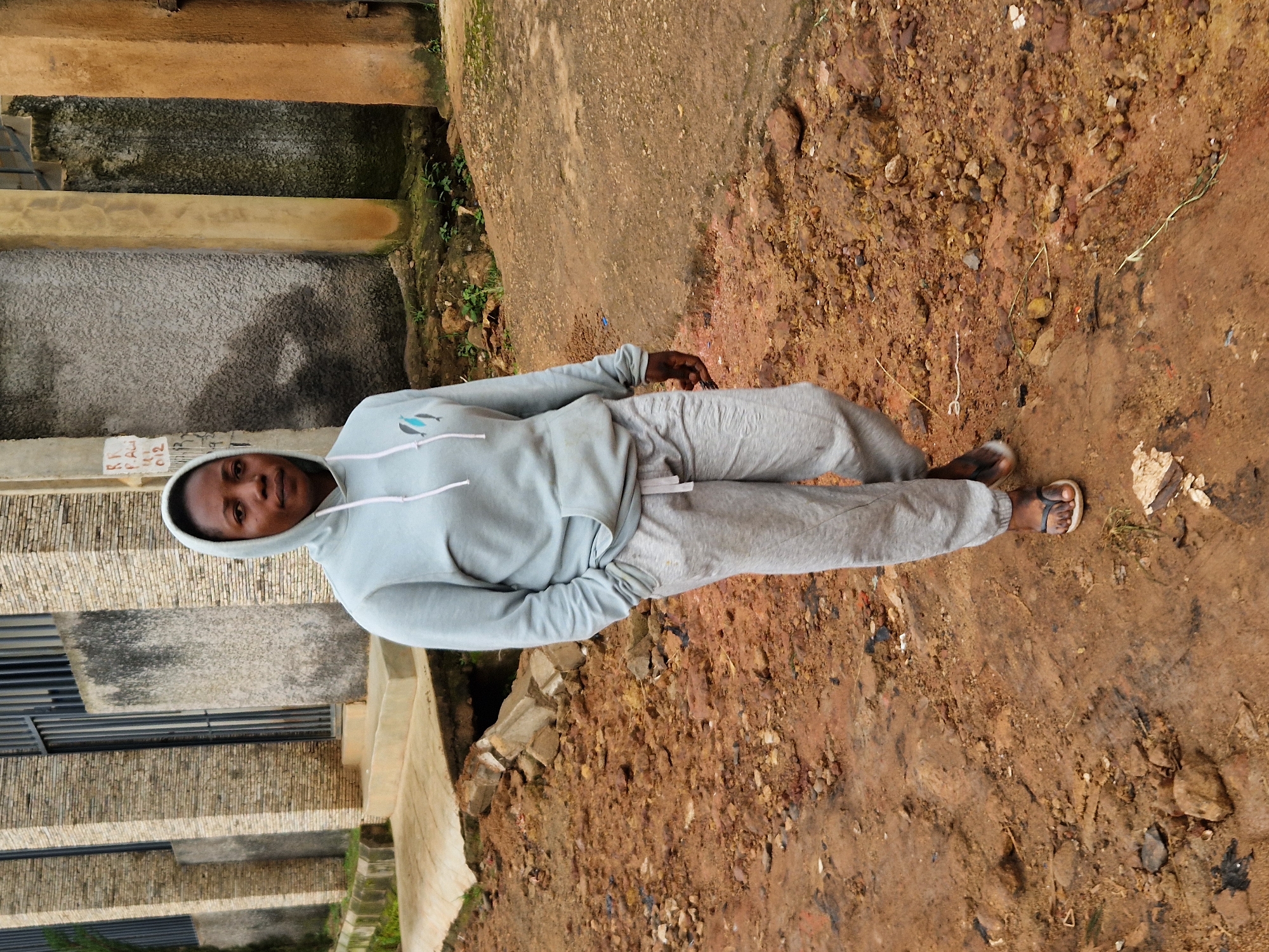 A full-body, eye-level outdoor shot depicts a Black woman with a medium skin tone and dark hair, wearing a light blue hooded sweatshirt with a white drawstring and light gray sweatpants. Her hands are tucked into the front pocket of her sweatshirt, and she is wearing dark flip-flops with white soles. She is walking from left to right across a dirt and gravel path. The background shows a textured, light gray wall on the left, a building with a barred window, and various architectural elements including concrete columns and a brown plastered wall. The ground is covered in reddish-brown dirt, rocks, and some debris. The scene appears to be located in the city of Kabong, Nigeria.