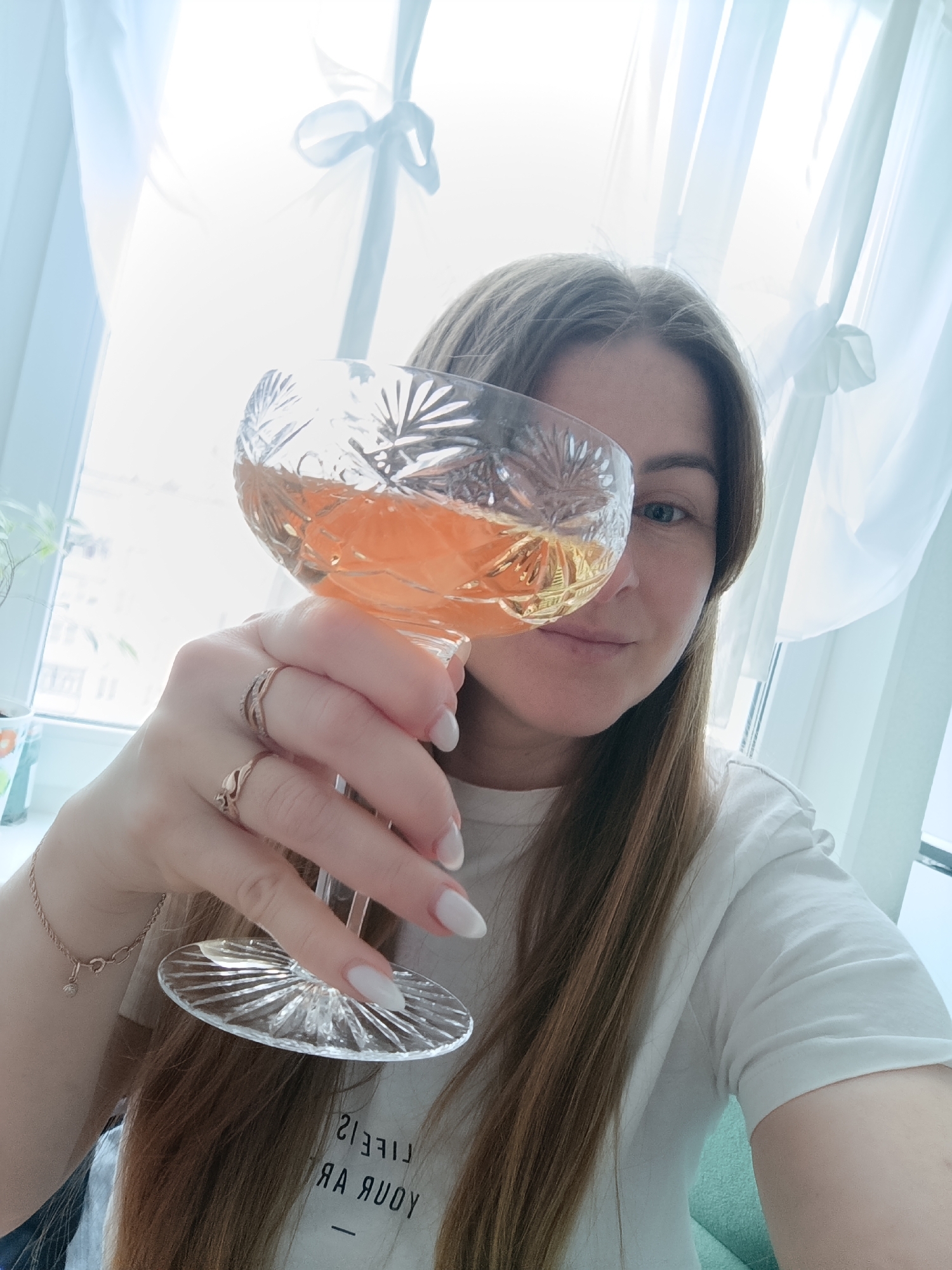 A woman with light brown hair is holding a glass of amber-colored liquid up to the camera. She has fair skin, a ring on her ring finger, and white nail polish. Her shirt is white and has some letters on it. The background consists of a window with white curtains and light streaming through it. The location is Unknown, Unknown.