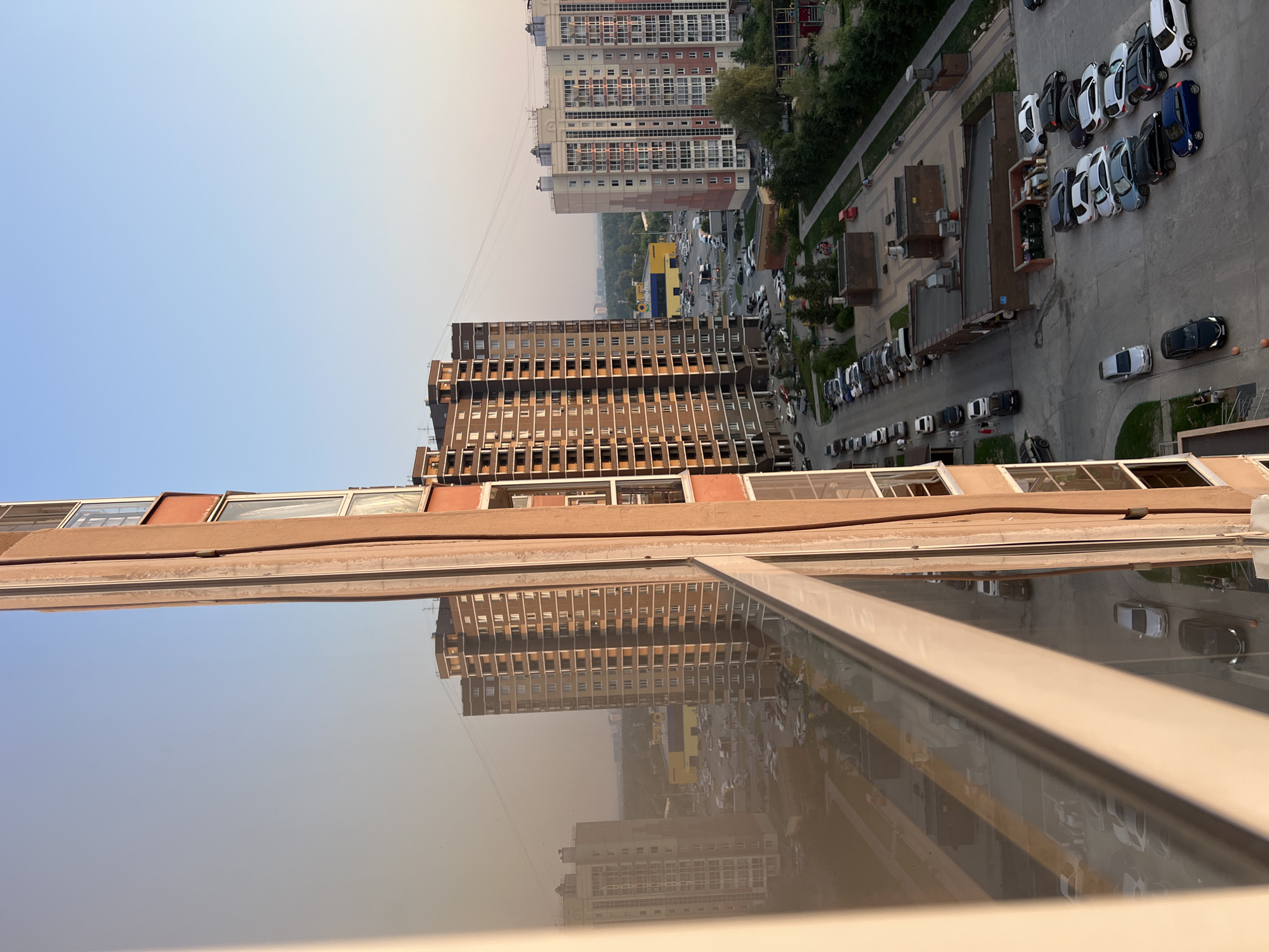 The image is a view from a high-rise building in Novosibirsk, Russia, overlooking a residential area. In the foreground, the reflection of a building is visible in a window. The main focus is on two tall residential buildings, one brown and one a lighter color with pink accents. A large yellow building with a blue and yellow sign is visible in the distance, likely a commercial establishment. The area is characterized by a network of roads and parking lots filled with various cars, including sedans, SUVs, and hatchbacks. Trees and green spaces are interspersed throughout the urban landscape. The sky is clear and blue, suggesting daytime. There are a few people visible walking on the sidewalks and in parking lots. The overall scene depicts a typical modern residential district in a Russian city.