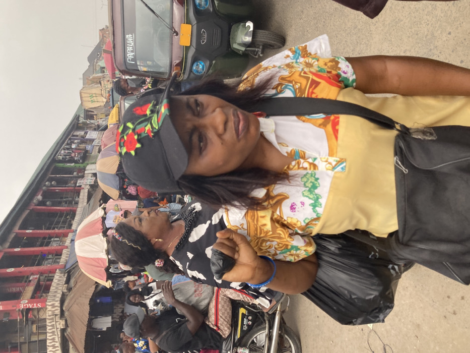 The image depicts a crowded outdoor market street in Aba, Nigeria, under an overcast sky. In the immediate foreground, an adult female of African descent is shown from the chest up, facing forward with a neutral to slightly focused expression. She wears a black baseball cap adorned with red rose embroidery and green leaf accents, a collared short-sleeved shirt with a plain yellow lower section, and a white upper section featuring yellow, orange, and green ornate patterns. A black shoulder bag is worn across her body, and she grasps a large black plastic bag in her right hand, revealing two blue bracelets on her wrist.
To her left, in the midground, an adult female of African descent with dark hair styled with a decorative headband wears a black and white patterned dress, a multi-strand black beaded necklace, and gold hoop earrings. She is looking towards the right. Further left, an adult male, also of African descent, wearing a black baseball cap and t-shirt, sits astride a silver motorcycle, holding a white plastic bag. The motorcycle's seat appears to have a patterned, possibly designer-branded, covering.
The background is densely populated with numerous individuals, market stalls displaying goods, and covered by various umbrellas, including a prominent red and white striped one. A multi-story building with distinctive red vertical structural elements and signage, including "STAGE" and "ARRIA," forms part of the background architecture. A green and black motorized tricycle, labeled "PAPAUWA" on its front, is visible on the right side of the midground. The ground surface is an asphalt or packed earth road, indicating active commerce and pedestrian traffic.