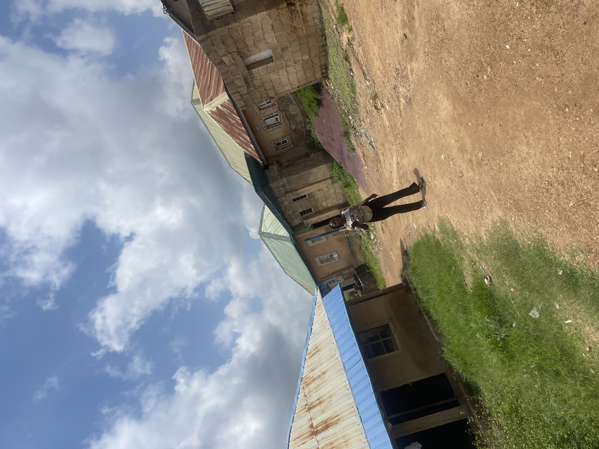 A woman stands in the middle of a dirt path, her body angled slightly to the left. She is wearing a light-colored, short-sleeved shirt with a pattern and dark, flared pants. Her left hand is raised and extended forward, while her right arm is by her side. The path she stands on is a reddish-brown dirt, with scattered pebbles and some green grass growing along the edges.
Behind her, a row of buildings forms the background. The closest building on the left has a light-colored, corrugated metal roof with a bright blue trim. Next to it, a building with a green roof appears behind and to the right. Further to the right, a larger, older-looking building is constructed with rough, light brown bricks. This building has several windows with decorative stone frames and a rusty, corrugated metal roof. To the far right, another section of a brick building is visible. The overall environment suggests a rural or semi-urban setting, possibly a residential area in Nigeria. The sky above is blue with scattered white clouds. The image is captured from a slightly low angle, looking upwards towards the sky and the buildings.