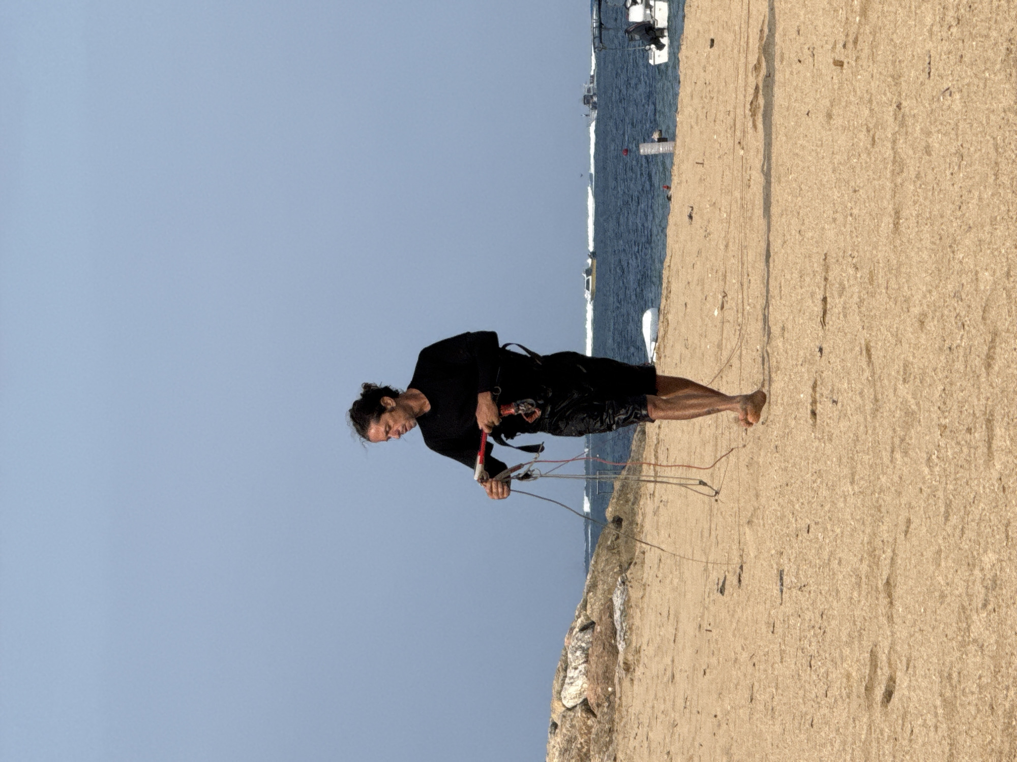 A man walks along a sandy beach in Denpasar, Indonesia, holding kiteboarding equipment. He is wearing a black long-sleeved shirt and black shorts. The beach is light brown with some rocks on the left side. The ocean is blue with several boats visible in the distance. The sky is a clear, pale blue.