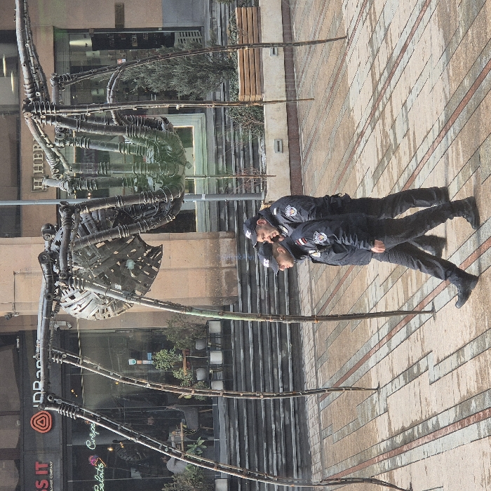 An outdoor urban scene in Yerevan, Armenia, specifically on Northern Avenue, features a large, dark-grey metallic spider sculpture standing on a patterned paved walkway. The sculpture is constructed from various metal components, including tubes, plates, and springs, giving it an industrial appearance, and has eight long, slender legs.
In the foreground, two adult males, identified as security or law enforcement personnel, walk from left to right across the paved ground. Both wear dark blue or black uniforms consisting of jackets, trousers, and baseball caps bearing a badge. Shoulder patches are visible on their left arms. The pavement is composed of rectangular tiles in light and dark shades, appearing damp in areas.
Behind the spider sculpture and the individuals, a series of multi-story commercial buildings line the background. On the left, a building with extensive glass windows displays "IDBank" and "Gelato Crepe" signage, revealing an interior with tables and chairs. Further right, "H HERMES" is visible on another building facade. Steps lead up to the entrances of these establishments. Small evergreen shrubs are present in planters or garden beds behind the sculpture's legs. The scene is illuminated by natural daylight.