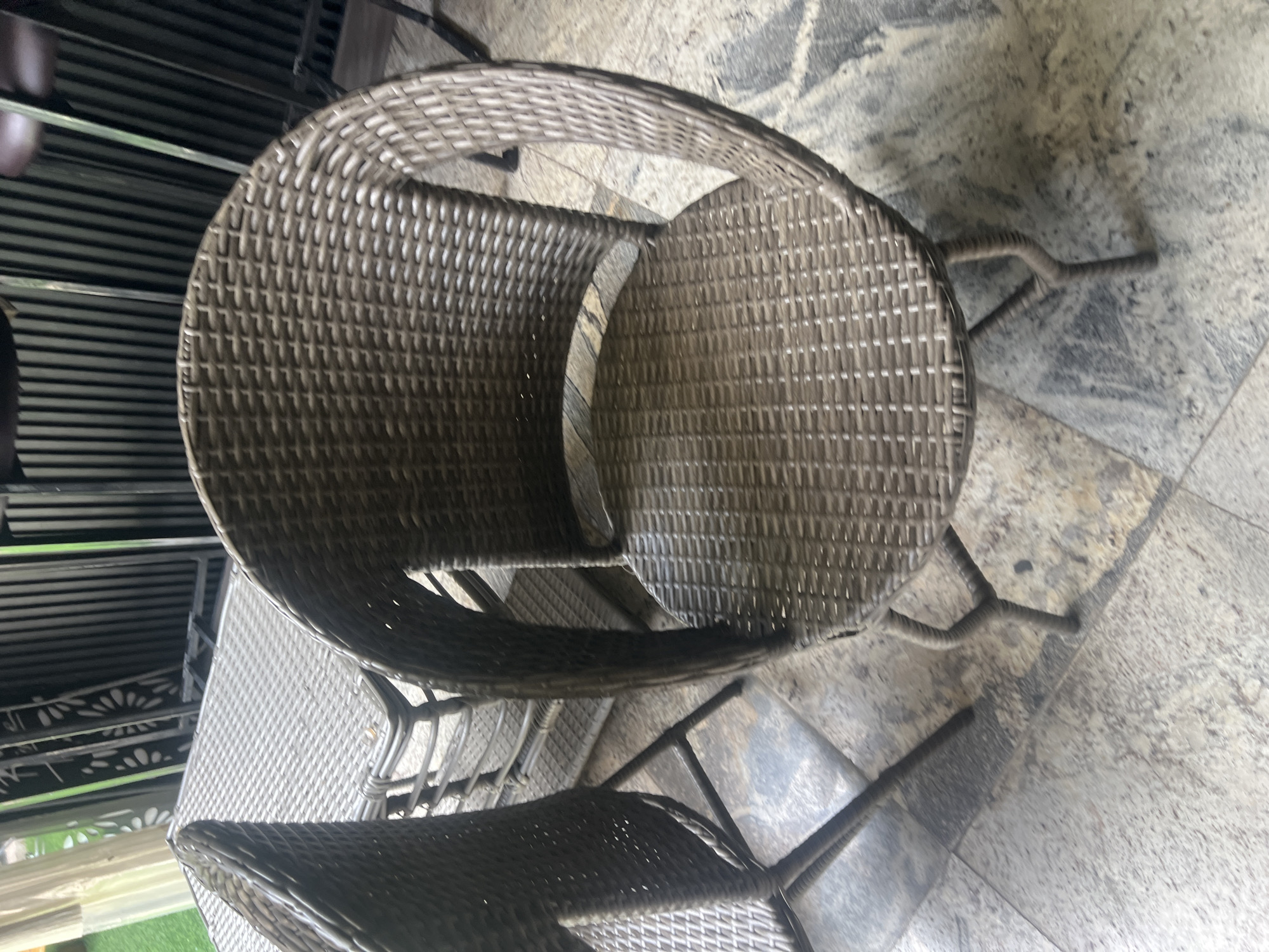 In Howayi, Nigeria, an outdoor patio area features a set of woven wicker furniture. The foreground is dominated by a brown wicker chair with a rounded back and a woven seat. The chair has four legs, with two visible in the image, designed in a slightly curved, ridged style. To the left, partially visible, is another matching wicker chair, and further back, a wicker table with a patterned surface is present. The furniture is situated on a floor paved with large, grey and beige stone tiles. In the background, a dark, slatted metal structure stands, possibly a fence or partition, with hints of greenery beyond. The scene is lit by natural light, suggesting an outdoor setting during the day. There are no people or animals visible in the image.