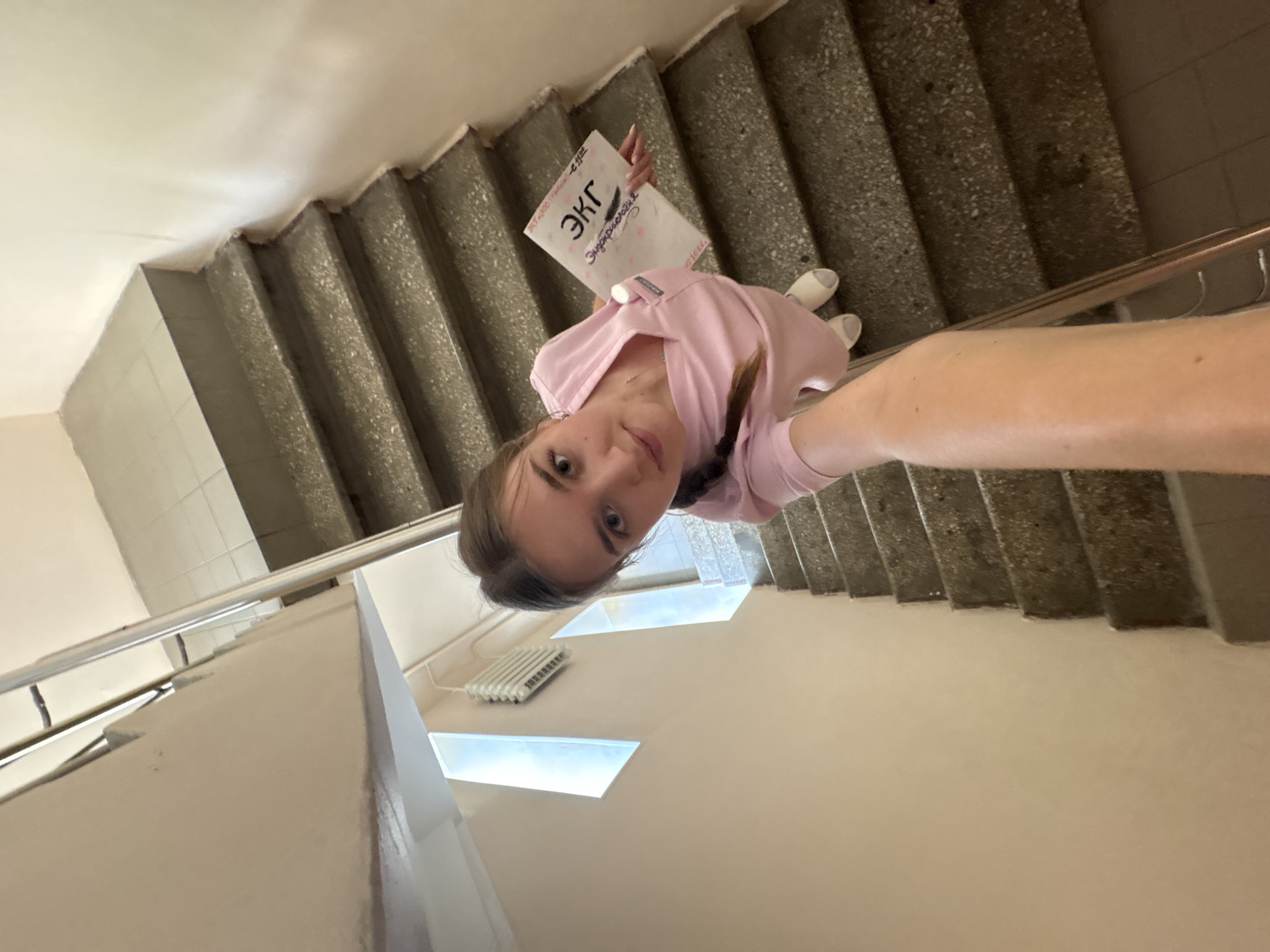 A young woman in a pink scrub suit stands on a concrete staircase, holding a document with Russian text. The staircase has metal railings and is lit by natural light from windows on the wall. The woman is looking at the camera with a slight smile. The background shows a tiled wall and a radiator. The scene appears to be in a public building or institution, possibly a hospital or school, given the attire of the woman and the document she is holding, which seems to be related to medical studies or practice. The specific location in the city of Perm, Russia, is not discernible from the visual information present in the image.