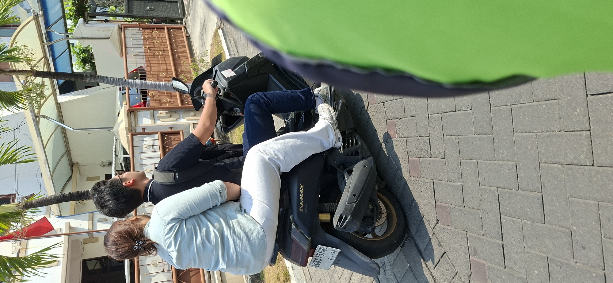 In Surabaya, Indonesia, two individuals are riding on a black Yamaha NMAX motorcycle on a paved sidewalk. The person in the rear, wearing a light blue long-sleeved shirt and white pants, is facing away from the camera. The person in the front, wearing a black shirt and dark blue jeans, is looking to the right, with sunglasses on. The motorcycle has a license plate visible that reads "DK 5575 FB". The background features a building with a red flag and palm trees, as well as a brown ornate fence. A blurred green object is in the foreground on the right side of the image.