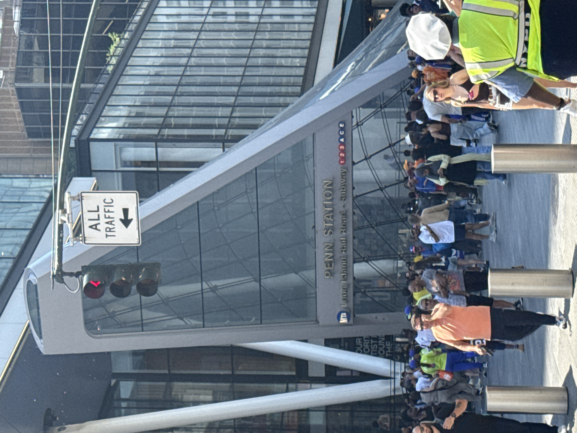 The image depicts a bustling outdoor scene in New York City, likely near Penn Station, as indicated by visible text. A crowd of people, diverse in age and attire, are gathered outdoors. Some are looking towards the camera, while others are engaged in conversation or movement. The architecture suggests a modern urban environment, with a large glass-fronted building dominating the background.
A prominent sign reads "ALL TRAFFIC" with an arrow pointing down, suggesting a road or pedestrian pathway. A traffic light is visible, showing a red signal. The overall atmosphere appears to be that of a busy public space, possibly a transportation hub or a popular gathering spot. The people's expressions are varied, with some appearing relaxed and others more preoccupied. The lighting suggests it's daytime, with sunlight casting shadows, and the weather appears to be clear. The presence of a person wearing a bright yellow safety vest might indicate some form of event management or construction work nearby. The various text elements, including "PENN STATION," "Long Island Rail Road," and subway line identifiers, confirm the location as a major transit point in New York.