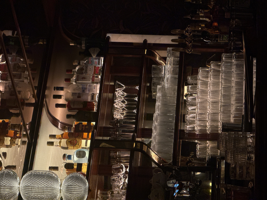 The photo captures a dimly lit bar setting. Shelves display an array of liquor bottles. Stacks of clear glasses are neatly arranged, alongside inverted martini glasses on a separate shelf. Ornate spherical decorations sit at the bottom of the shelf. A curved brass accent is visible, and an espresso machine is located in the bottom right. The lighting is low, casting shadows throughout the scene.