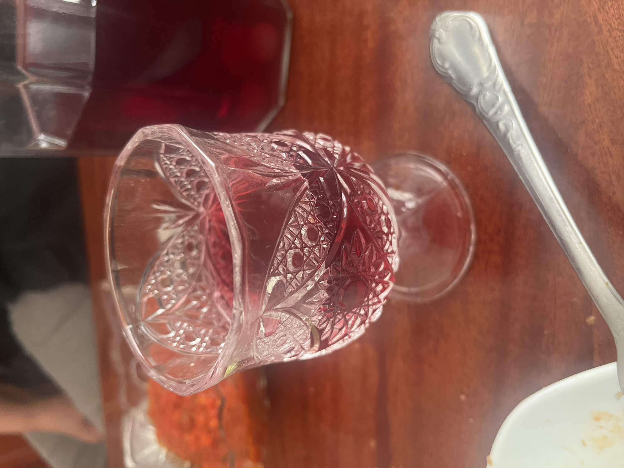 A high-angle shot shows a cut-glass wine glass filled with a red liquid, most likely wine, on a brown wooden table. The glass has an intricate, faceted design. To the right of the glass is a silver spoon resting beside a white plate, possibly containing food remnants. In the upper left corner, a glass bottle filled with a similar red liquid is visible. The composition suggests an indoor setting, perhaps a dining area. There is no discernible information to infer any association with the city of Yeghegnadzor, Armenia.