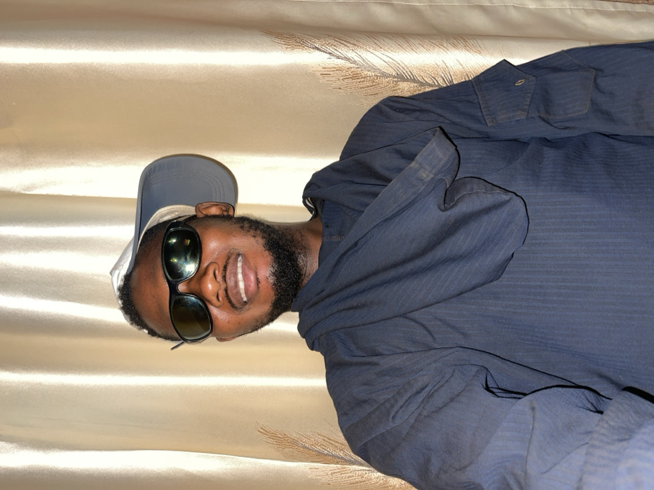 A man in sunglasses and a baseball cap smiles at the camera. He has a dark beard and wears a dark blue collared shirt. The background is a soft, beige fabric with a subtle sheen and a feather design. The image is taken at an angle, with the man positioned sideways relative to the frame.