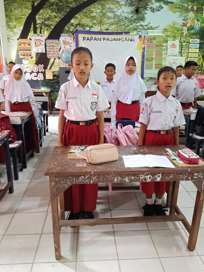 A classroom scene shows Indonesian elementary school students in uniform standing and seemingly reciting or singing. They wear white shirts with red skirts or pants, with some girls wearing headscarves. A display board is behind them. Desks are in front of the students, with open notebooks and pencil cases on some. The students appear to be focused and attentive.