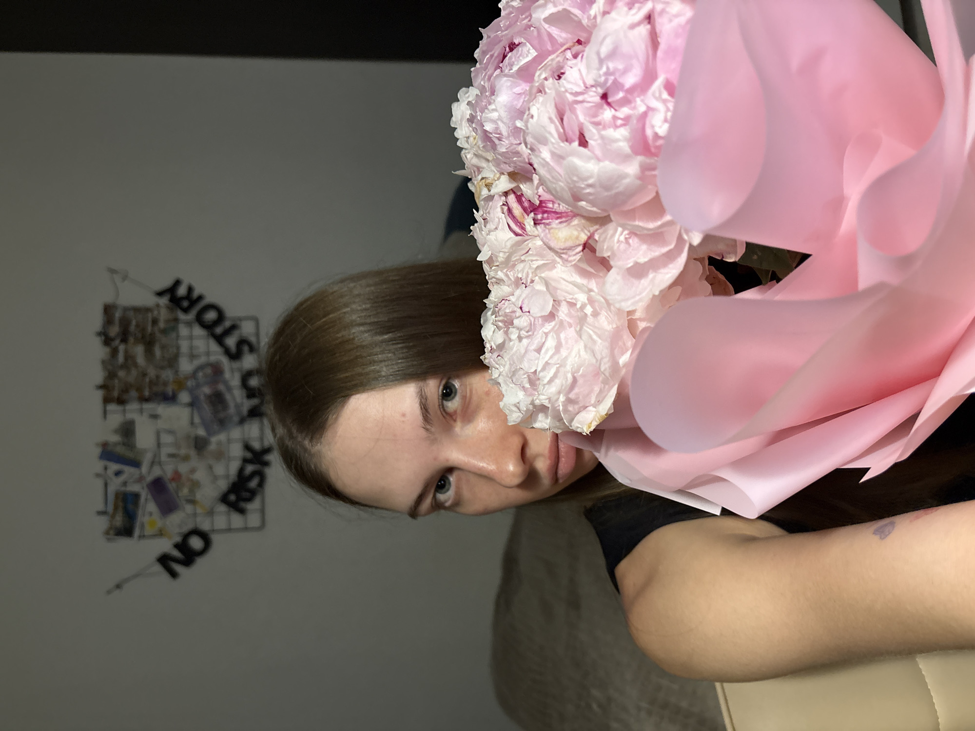 A young woman with long, brown hair and blue eyes is holding a bouquet of pink peonies, partially obscuring her face. She is wearing a black top. In the background, a wire grid is mounted on a light-colored wall, decorated with various pictures and text. The text on the grid reads "NO RISK NO STORY" in black letters. The overall setting appears to be indoors, possibly a bedroom, with a beige chair visible in the foreground. The lighting is dim, suggesting it is nighttime or the room is not well-lit. The specific location within the city of Perm, Russia, cannot be determined from the provided image.