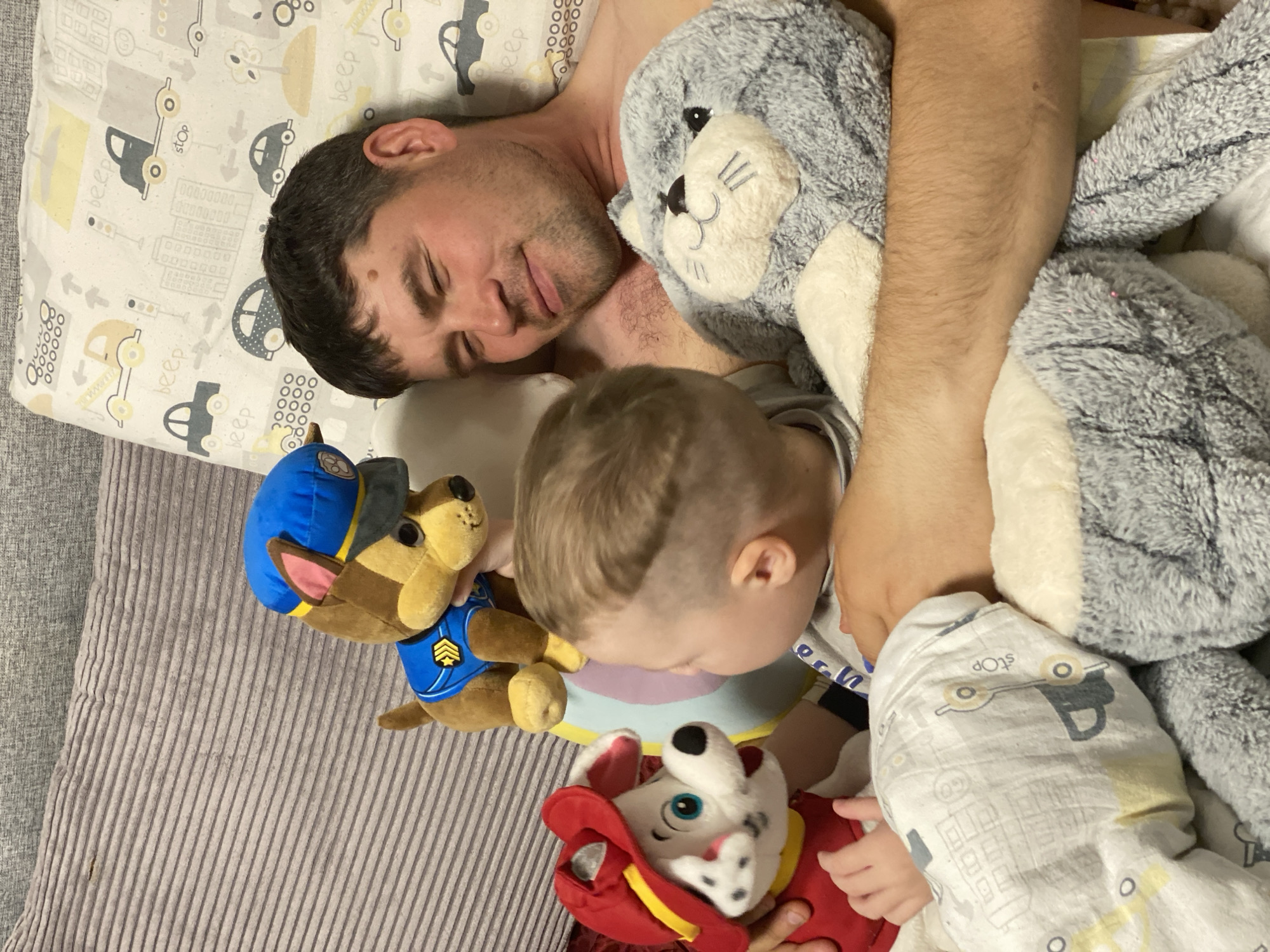 This indoor image captures a tender and peaceful moment between a father and his young son, likely in a bedroom in Sochi, Russia. The adult male, with short dark hair and some stubble, is seen sleeping or resting on his side with closed eyes. He is shirtless and appears to be spooning a large, fluffy gray seal or sea lion plush toy.
Beside him, his young son, whose back of the head and partial face are visible, is also resting. The child has light-colored hair and is holding two specific plush toys from the popular Paw Patrol series: Chase, the police dog, and Marshall, the fire dog. The child's hand is visible, gently grasping the Marshall toy. He is partially covered by a white swaddle blanket featuring a playful pattern of cars, roads, and traffic signs in muted yellow, gray, and black, identical to the main bedsheet.
The bedding includes this light-colored sheet with various road-themed elements. Visible text on the fabric includes "stop," "beep," and "GO," alongside numerical patterns such as "000," "0000000," and "00000000." A textured, light gray ribbed blanket or cushion is also visible to the side. The overall scene conveys a strong sense of comfort, warmth, and tranquility, suggesting a naptime or bedtime shared by parent and child.