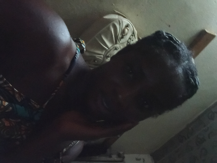 A woman with dark skin, braided hair, and a colorful top rests her head on her hand, looking at the camera. The lighting is dim. Behind her is a cream-colored cushion on a couch, and part of a wall is visible in the background. The image appears to be taken at a tilted angle.