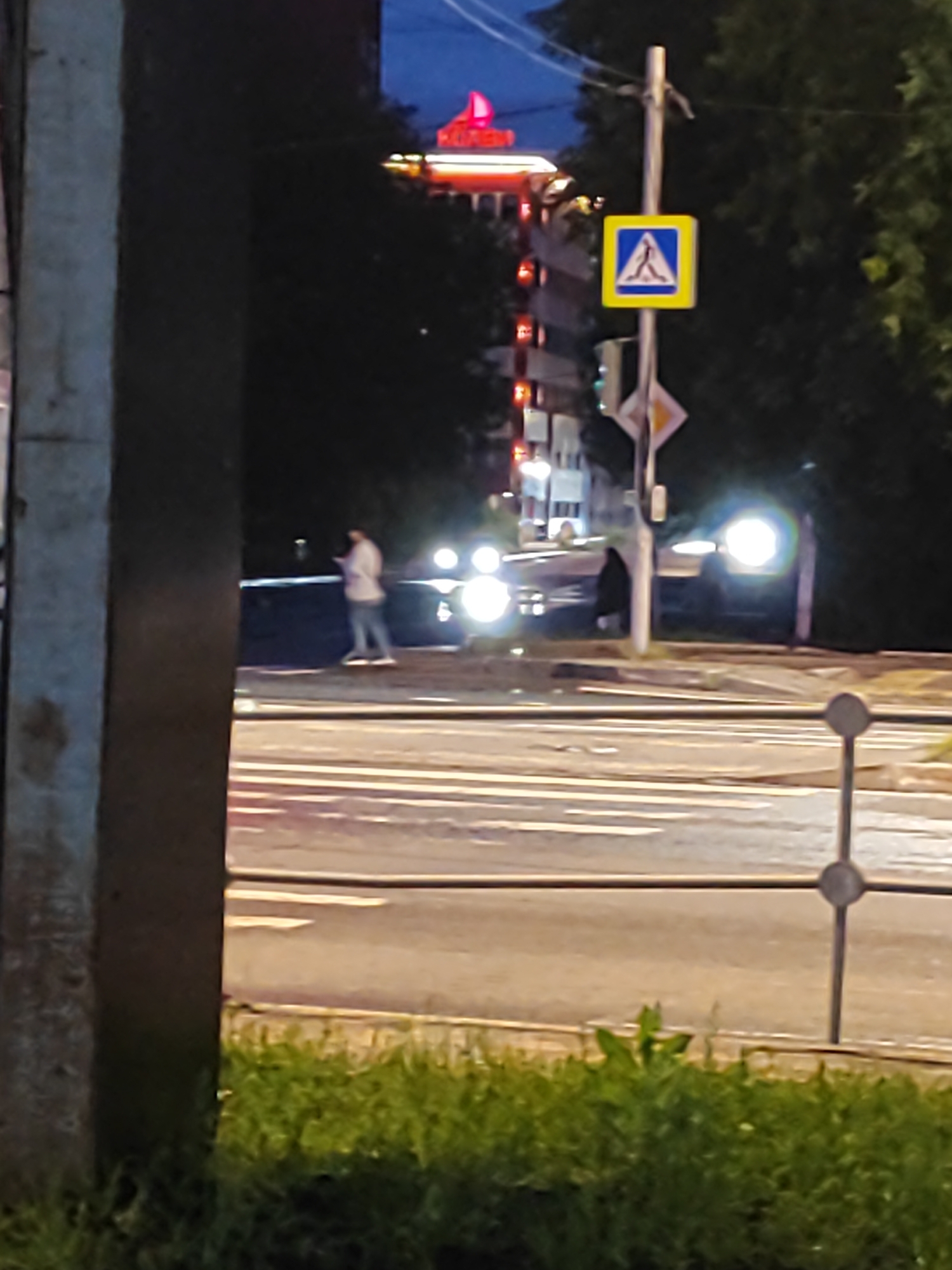 The image depicts a nighttime street scene. In the foreground, a dark, textured vertical object, possibly a utility pole or a building facade, occupies the left side of the frame. To the right, a pedestrian crossing with white stripes is visible on a paved road. A figure, appearing to be a person, is walking on the street, facing away from the viewer, and holding a phone. Bright headlights from unseen vehicles illuminate the road. A traffic sign, a yellow square with a blue border and a white pedestrian symbol, is mounted on a pole. Behind the street, trees with dark foliage line the background. In the distance, a tall building with illuminated red and yellow lights is visible against a dark blue sky. The scene appears to be in an urban environment.