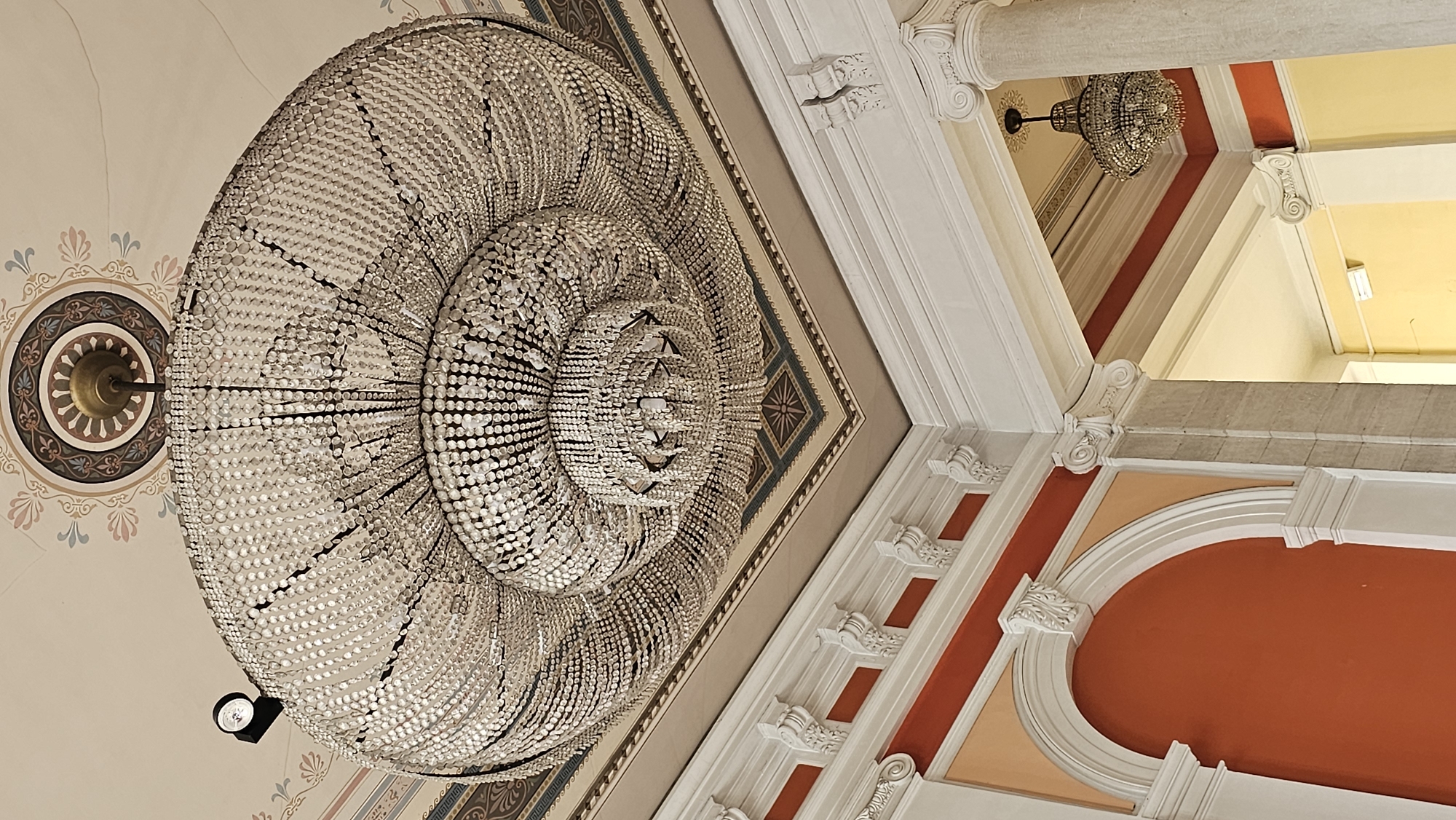The image depicts an ornate interior, likely a historic building, in Varna, Bulgaria. A large, multi-tiered crystal chandelier dominates the view, suspended from a high ceiling with decorative moldings and painted patterns. Another smaller chandelier is visible in the background. The architectural details include white pillars, red and peach painted walls, and a patterned ceiling border. No people are visible in the frame.