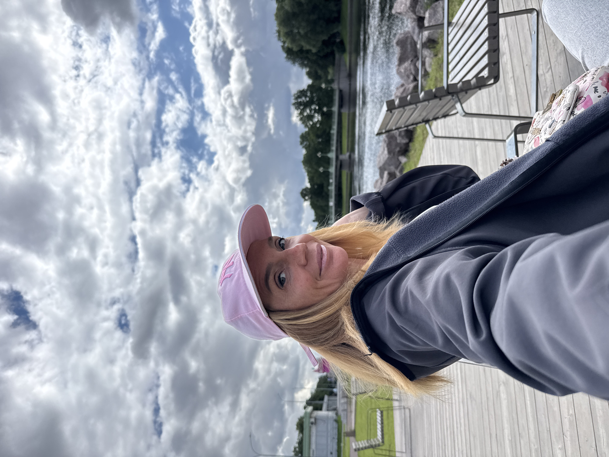 A woman with blonde hair and a pink baseball cap smiles at the camera while taking a selfie. She is wearing a dark gray jacket with a gray fleece lining visible at the collar. The background shows a cloudy sky, a body of water, a bridge, trees, and a boardwalk with benches. The location is likely near the Neva River embankment in Saint Petersburg, Russia, given the architectural style of the bridge and the presence of the river. The scene appears to be set in a park or public recreational area.