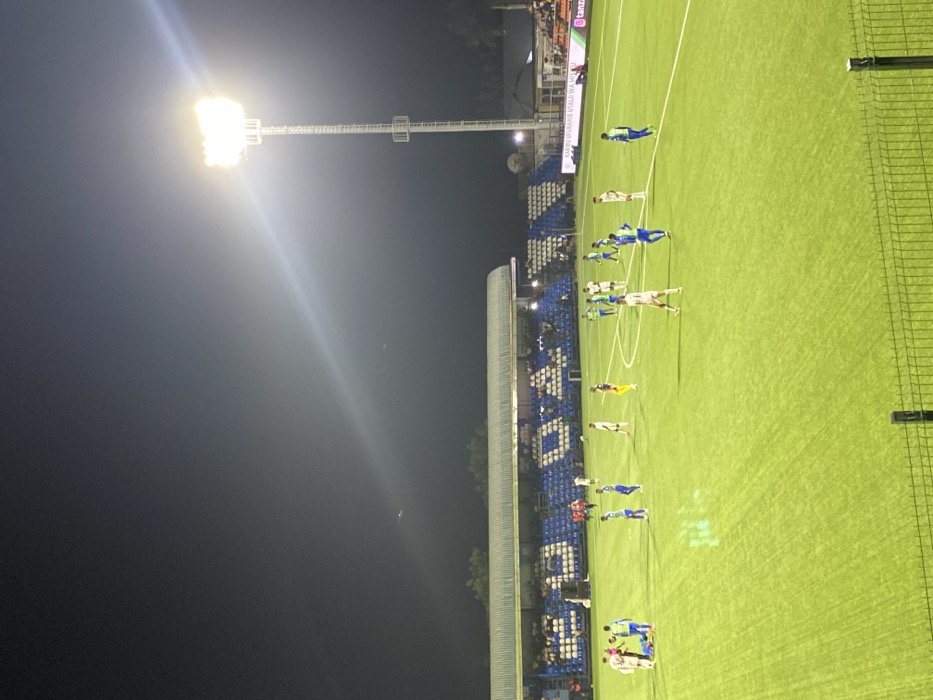 The image shows a soccer field at night, illuminated by a single bright spotlight. The field is green, with white lines marking the pitch. A group of about 10 soccer players wearing blue and white uniforms are standing on the field, seemingly preparing for a match. The players are positioned on the right side of the image, in front of the stadium stands, which are full of spectators. The stands are a vibrant blue color and have a white covering. The image was taken from the stands and the camera is positioned directly behind the players. In the distance, beyond the stands, there are trees and other structures, likely part of the stadium complex. The night sky is dark, with a few stars visible in the background. The location is in Dar es Salaam, Tanzania, at coordinates -6.97 latitude and 39.223 longitude.