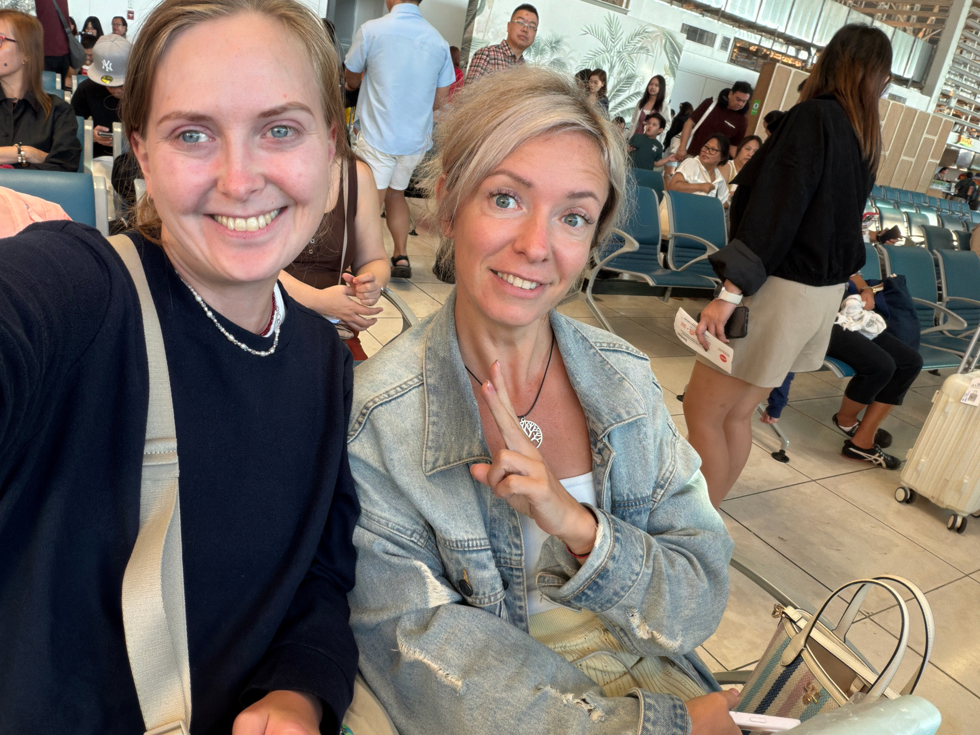 In Pasay City, Philippines, two women are taking a selfie inside an airport terminal. The woman on the left, with blonde hair, wears a dark blue long-sleeved shirt and a pearl necklace. The woman on the right, also with blonde hair, sports a distressed denim jacket over a white top and a necklace with a tree pendant. Both are seated in what appear to be airport waiting area chairs, with other travelers and luggage visible in the background. The environment is brightly lit, suggesting it is daytime, and the setting is characteristic of an indoor public transportation hub.