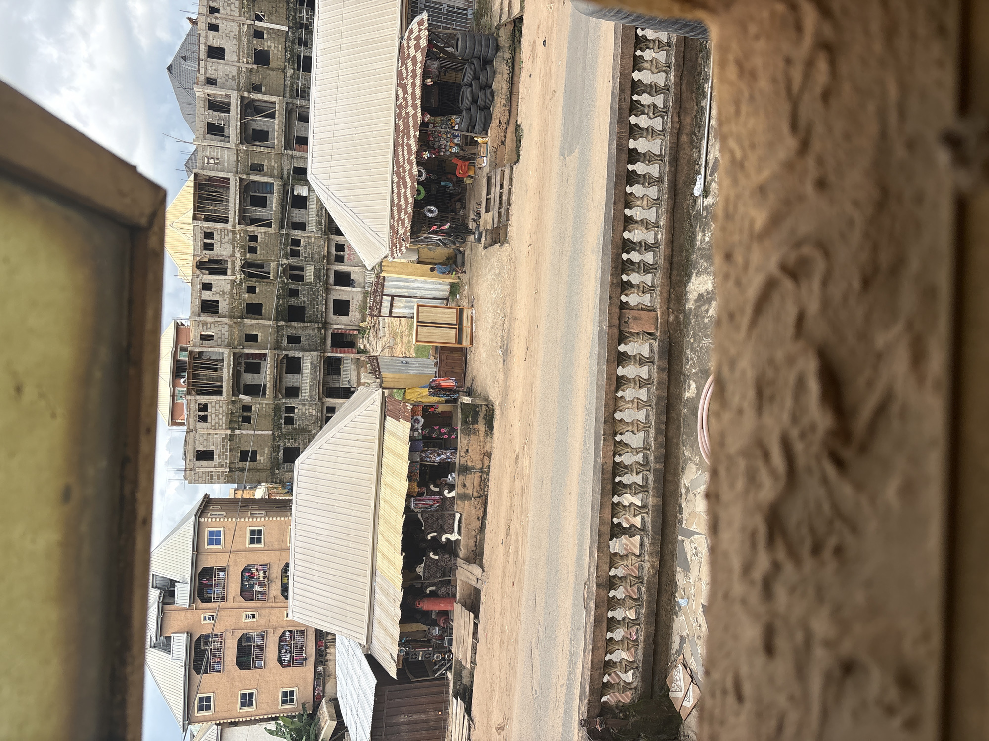 This image depicts a street scene in Nkpor, Nigeria, characterized by a mix of residential buildings and small commercial stalls. In the background, several multi-story buildings are visible, some appearing to be under construction with exposed concrete blocks and scaffolding. One building is complete, featuring multiple balconies with laundry hanging out to dry.
In the foreground, two open-air stalls with corrugated metal roofs line a dirt road. The stall on the left displays various items, including what appear to be tires, a red gas cylinder, and shelves stocked with merchandise. The stall on the right also exhibits tires stacked on the ground, along with racks of goods, a red chair, and some hanging fabrics or garments. A person, a young boy, is seen walking away from the stall on the right.
The road is unpaved and dusty, with a decorative concrete balustrade running along the edge of a raised area in the foreground. The environment appears to be a bustling urban setting with signs of ongoing development and commerce. The lighting suggests it is daytime, with a cloudy sky.