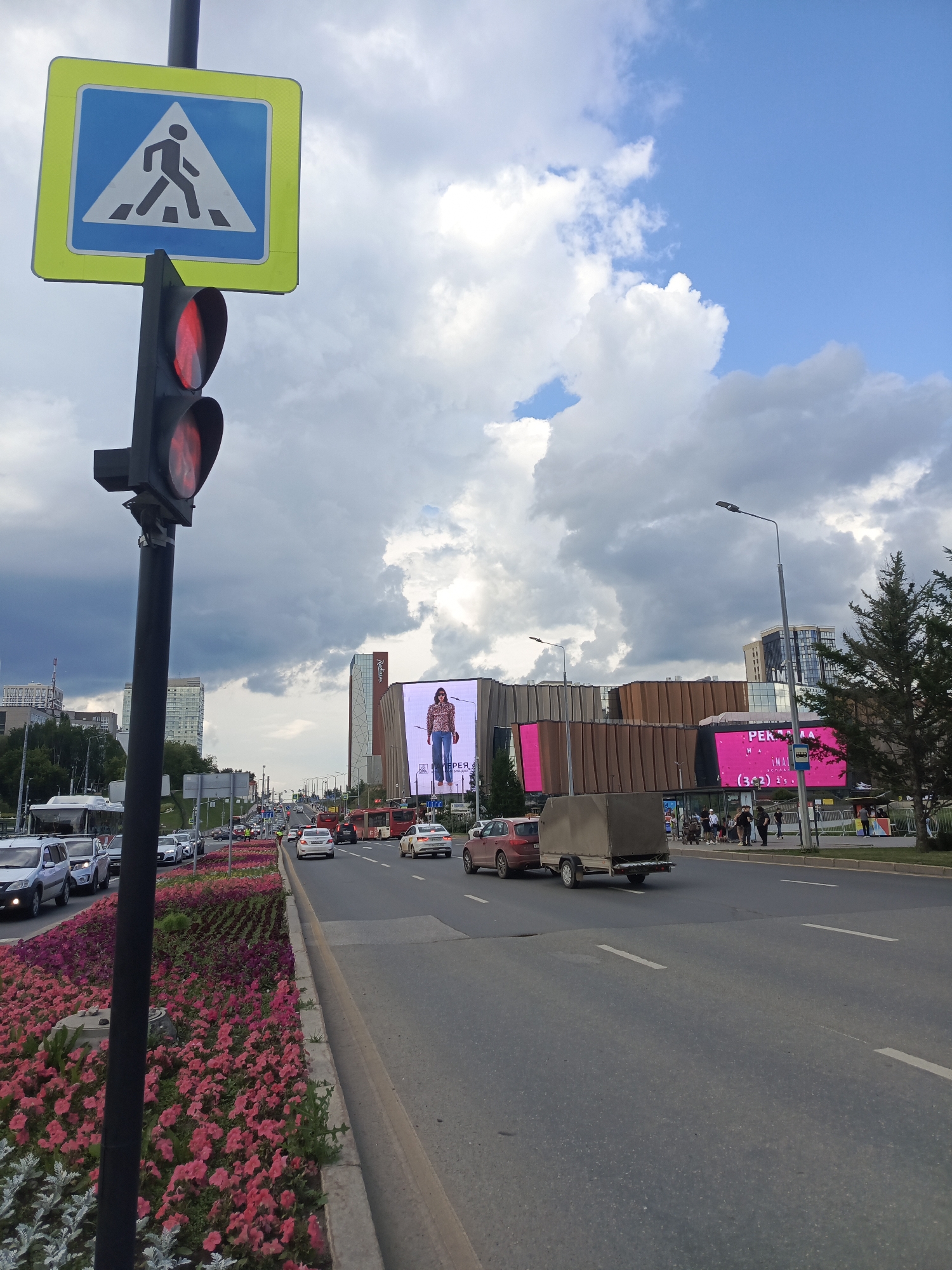 The image depicts a street scene in Perm, Russia, on a partly cloudy day. A pedestrian crossing sign and a traffic light showing red are prominently displayed in the foreground, positioned near a busy road. The road is lined with vibrant pink and purple flowers, creating a decorative median. Several cars and a bus are visible on the road, indicating traffic. In the background, a large modern building with several screens displaying advertisements stands out. One screen features a model in casual clothing, and another shows pink graphics with text. The architecture of the building suggests it is a shopping center or entertainment complex. People can be seen walking along the sidewalk in front of the building. The sky is a mix of blue and dramatic grey clouds, hinting at changing weather. The overall impression is an urban environment with commercial activity and traffic flow.