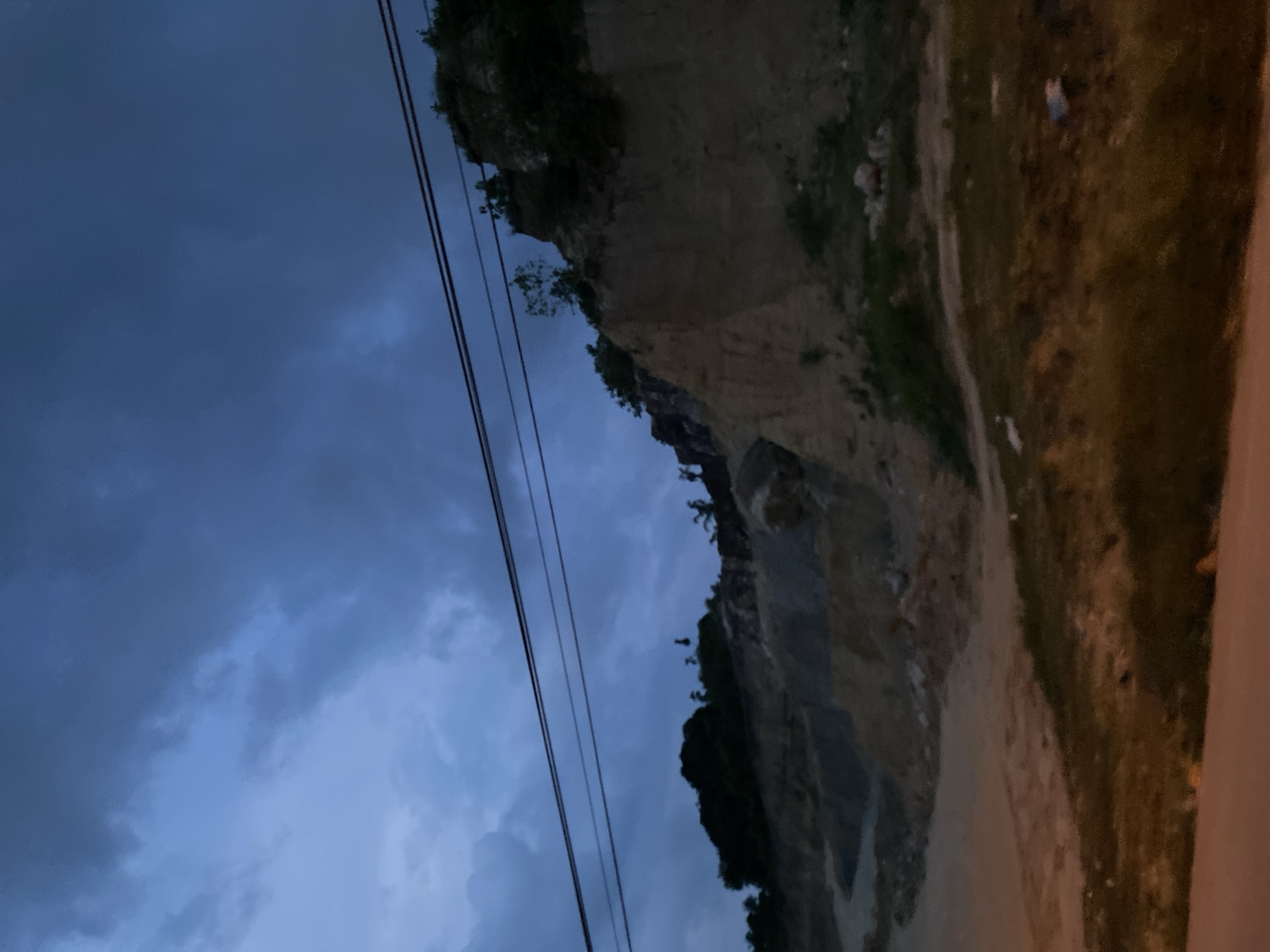 The image shows a roadside view in Lhokseumawe, Indonesia, at dusk or night. The sky is overcast with dark, heavy clouds. To the right is a high, rocky cliff face with sparse vegetation clinging to it. A dirt road runs along the base of the cliff. Power lines are visible in the foreground against the sky. The scene is dimly lit.