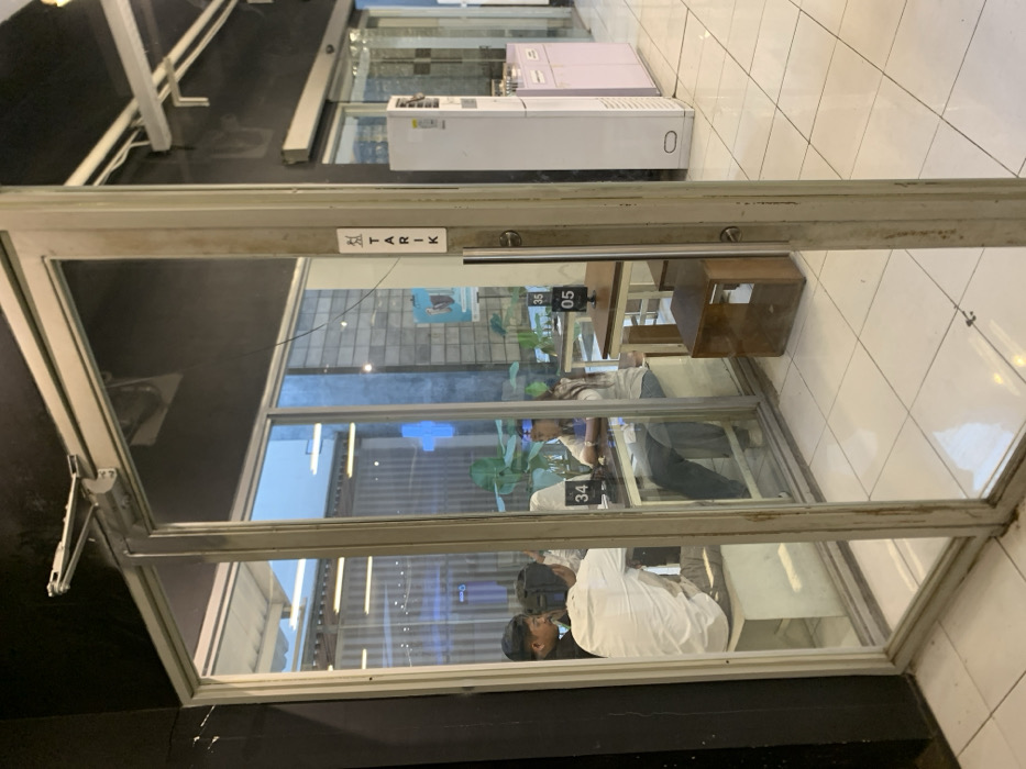 The image shows a view through a glass door, looking into a cafe in Malang, Indonesia, located at latitude -7.941 and longitude 112.608. There are four people seated at two tables, engaged in conversation. One man is wearing a white shirt, another man is wearing a white shirt, and a woman is wearing a white shirt. The tabletops are marked with numbers 34 and 35, indicating seating arrangements. There are also visible signs and other furniture. The cafe's floor is tiled in white. The environment is bright and well-lit. The door is partially open, with a sign that says "TARIK" visible on the door frame.
