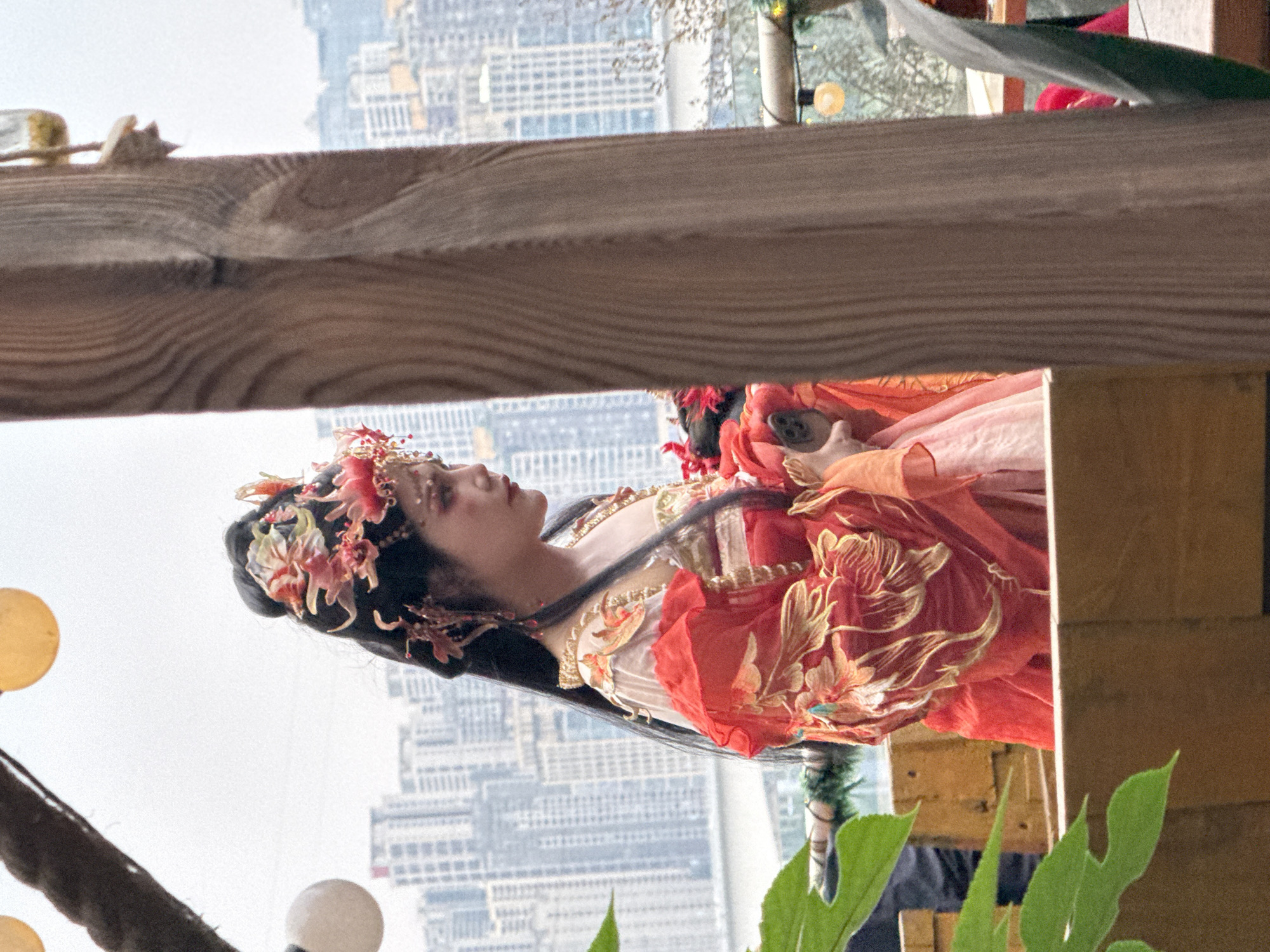 A medium shot depicts a person in traditional Chinese attire against the backdrop of an urban landscape. The person, likely female, has long black hair adorned with floral hair ornaments. She wears a layered, red and cream-colored garment with ornate gold embroidery. The attire suggests a historical or cultural performance context.
The background features tall, densely-packed buildings, typical of a modern city center. The buildings appear in the distance, slightly obscured by haze or atmospheric perspective. The location is consistent with the urban environment of Yuzhong District, China, considering its high-rise architecture and bustling city characteristics.
In the foreground, elements of what appears to be an outdoor set or display are visible, including wooden beams, foliage, and globe-shaped lights. The presence of these elements suggests the subject is posed or situated in a specific environment for a photograph or performance.