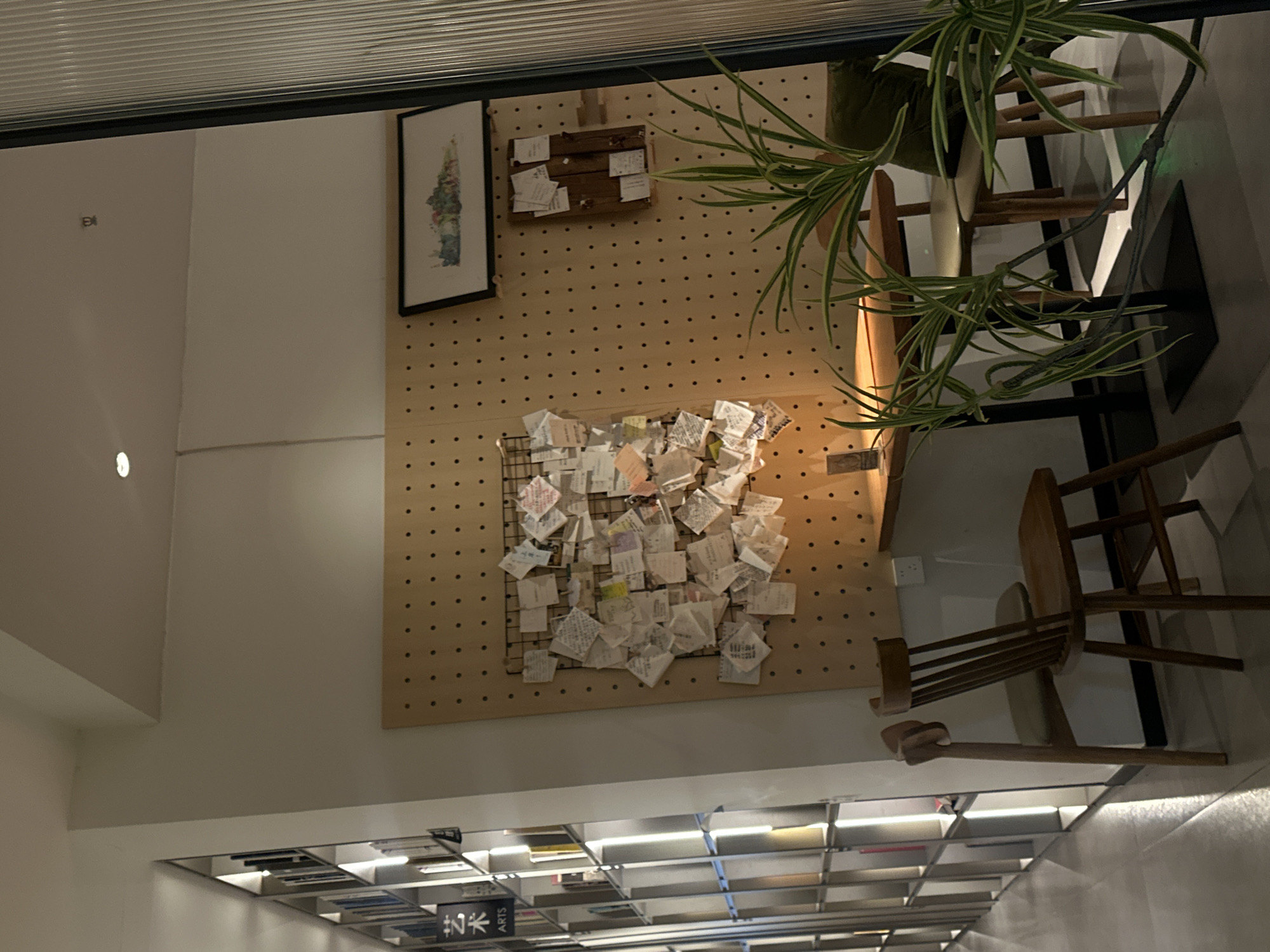 This image depicts an indoor commercial or public space, specifically a quiet corner within the Chongqing High-tech Zone, China. The scene is devoid of people. On the left, a tall, multi-shelved bookcase, illuminated vertically, displays numerous books; a black sign with white text "艺术 ARTS" is affixed to one shelf. The central background features a light-brown wooden pegboard wall. A large black wire grid is mounted on this pegboard, densely covered with multiple small, folded paper notes, indicative of a community message board. To the right, a smaller wooden plank board also holds a few pinned notes. Above these boards, a horizontally oriented framed artwork, likely a landscape painting, is hung. In the mid-ground, a wooden table with a black metal base is positioned, flanked by two wooden chairs; one chair has a light-colored cushion. A white electrical outlet is visible on the wall beneath the pegboard. A green potted plant with long, variegated leaves occupies the immediate foreground on the right, partially obscuring the table and chairs. The ceiling is white and features recessed spotlights. A vertical, translucent fluted glass panel defines the rightmost edge of the frame. The overall environment presents a clean, organized, and inviting atmosphere, potentially a cafe or reading area.