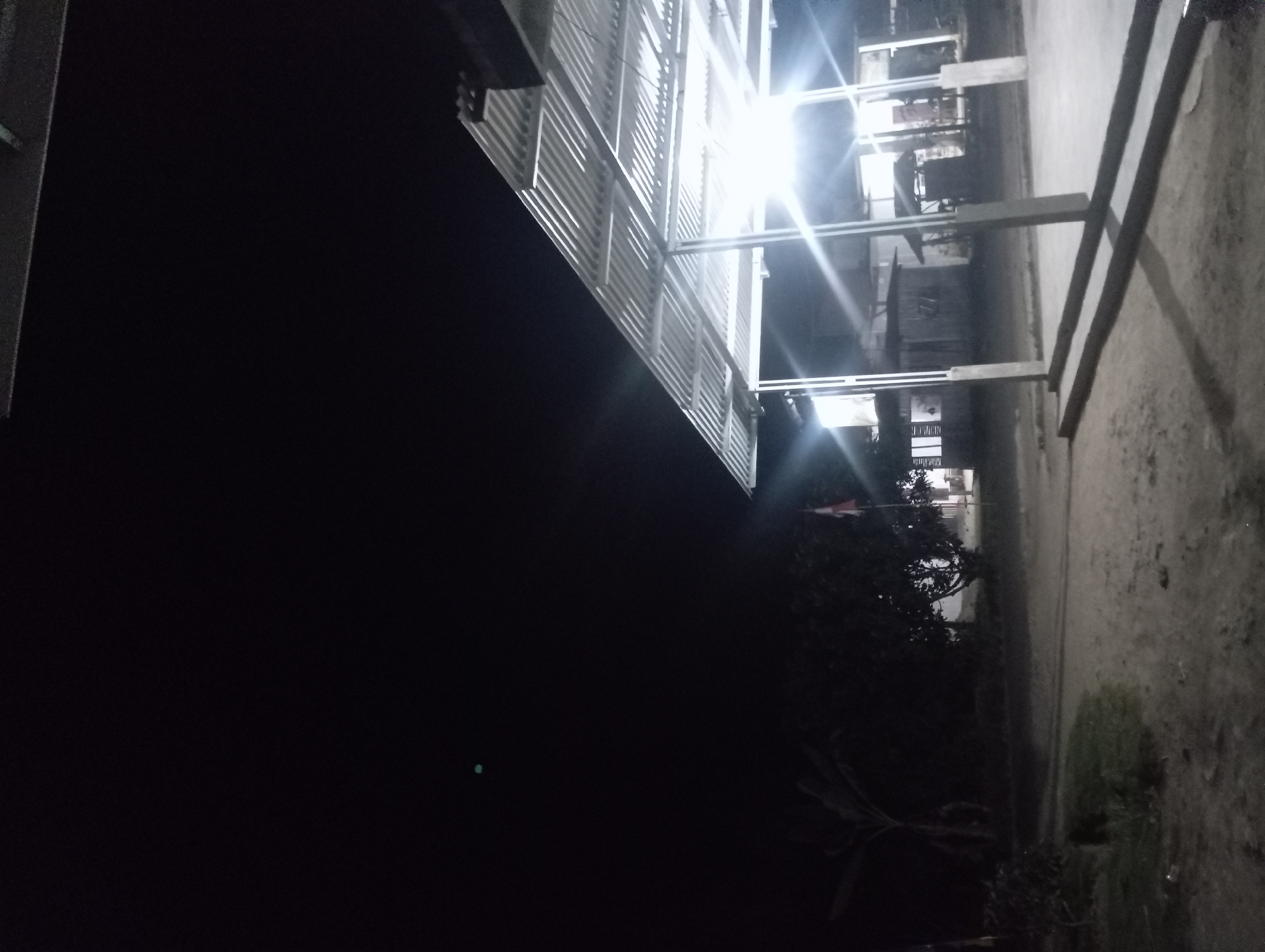 The image depicts an outdoor scene at night in Rejosari, Indonesia. A corrugated metal roof structure, supported by pillars, is illuminated by bright lights, casting long shadows on the sandy ground. In the background, several small buildings or shacks are visible, some with lights on. A flag, possibly the Indonesian flag given the location, is seen on a flagpole. Trees and foliage are present, contributing to a rural or semi-rural setting. The sky is dark, indicating nighttime. There are no people visible in the image.