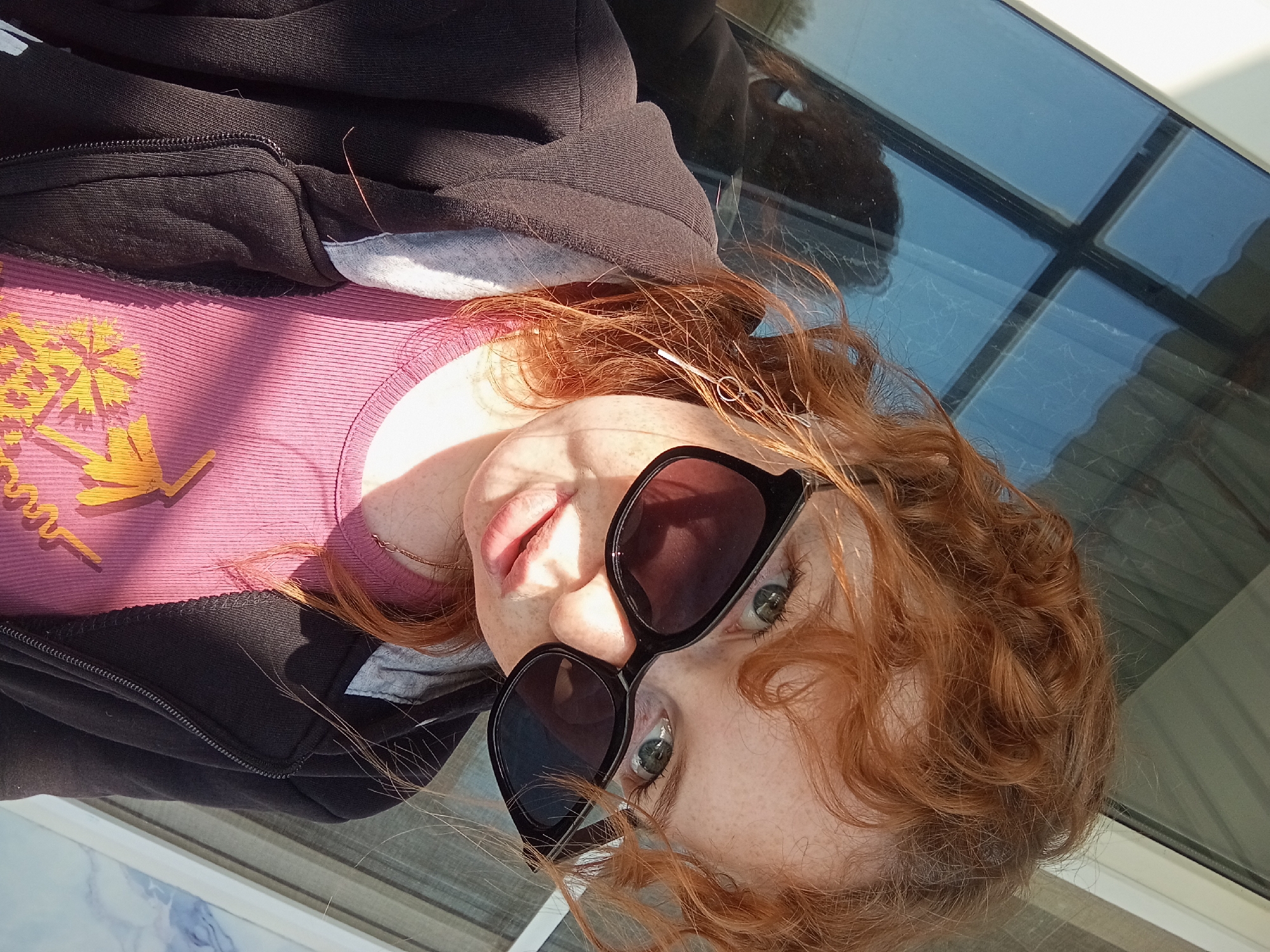 A selfie-style image shows a fair-skinned person with freckles, wearing dark sunglasses, a pink t-shirt with a yellow palm tree graphic, and a partially zipped-up black hooded sweatshirt. The person has red, curly hair and is positioned at an angle in the frame. The background includes a window with visible reflections. The location in Yurgamysh, Russia is unverifiable from the image. No specific actions or interactions are discernible.