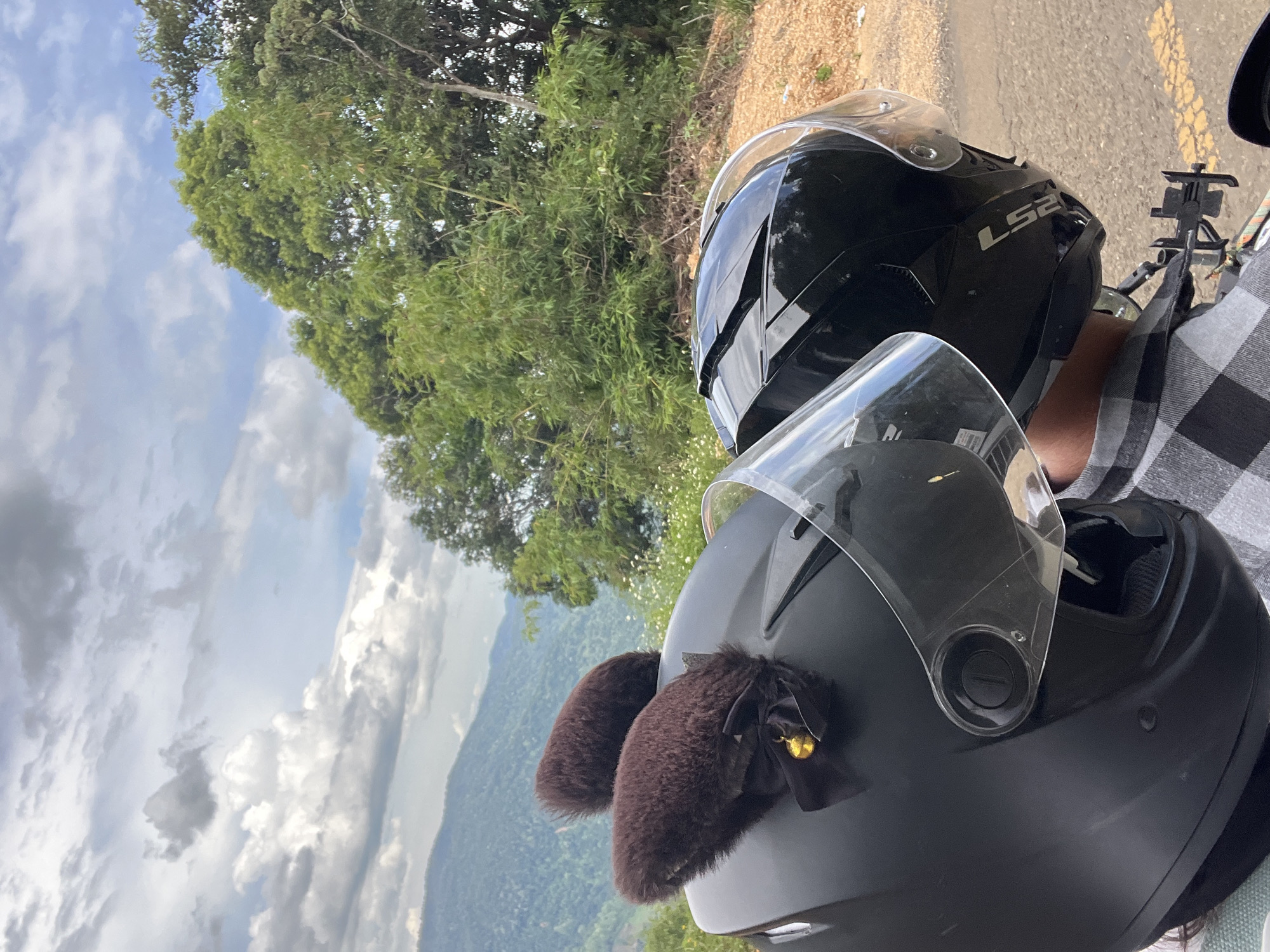 Two people wearing helmets are on a motorcycle, likely in Son Dien, Vietnam, based on the lush green hills and cloudy sky. The helmet in the foreground is black and decorated with brown furry "cat ears" and a small gold bell. The second helmet, to the right, is also black and has a clear visor. The background features rolling green hills covered in vegetation, with a dense cluster of trees to the right. The sky is partly cloudy with a mix of grey and white clouds. The ground appears to be a road with some loose gravel and a yellow line marking. The overall scene suggests a scenic ride through a rural or mountainous area.