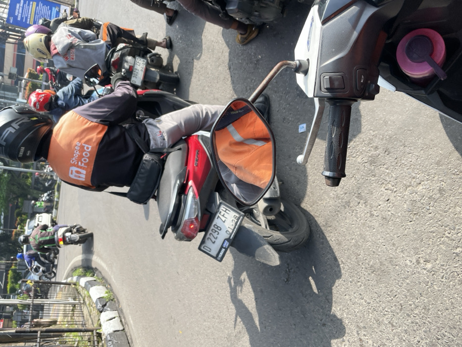 The image depicts a street scene in Bandung, Indonesia. The location is identified as being near the intersection of Jalan Ir. H. Juanda and Jalan Otto Iskandardinata based on the latitude and longitude coordinates. The scene is dominated by a black and white motorcycle in the foreground. The motorcycle is facing the street, and the handlebar is visible. The rearview mirror reflects an orange jacket worn by a person on a red motorcycle. The motorcycle has a license plate that reads "D 2298 ZFH." In the background, there are several other motorcycles and pedestrians. The street is paved and there is a sidewalk to the left of the frame. Several buildings can be seen in the background. The sky is partially obscured by tree branches, indicating that it is daytime and likely a partly cloudy day. The environment appears to be an urban area with busy traffic and multiple vehicles. The image captures a moment in the daily life of Bandung, showcasing the presence of motorcycles and pedestrian movement within the city.