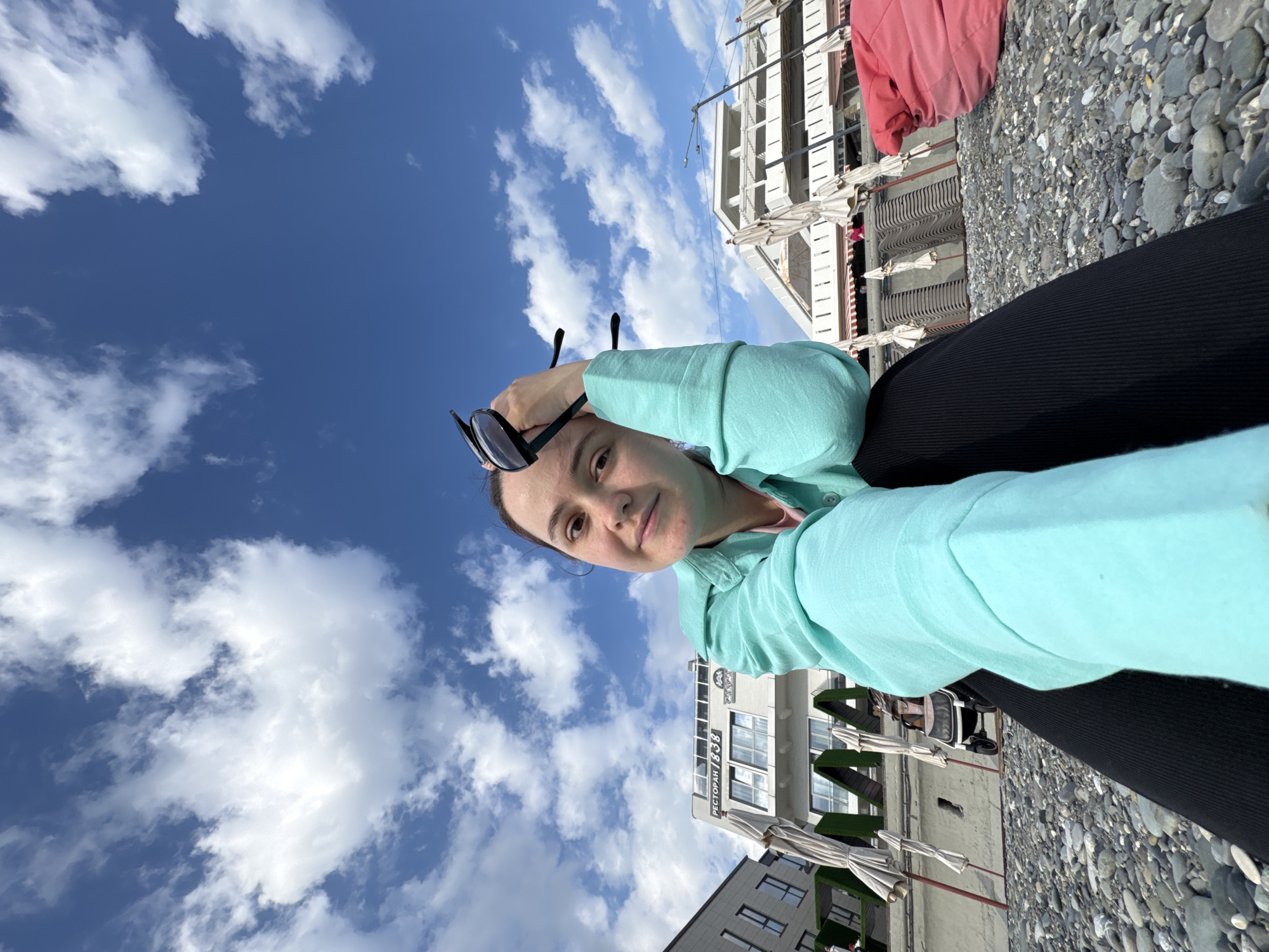 The image shows a woman taking a selfie on a beach in Sochi, Russia. She is wearing a light blue sweatshirt and black leggings. She has sunglasses propped on her head. Behind her are buildings, a pebble beach, and a partly cloudy blue sky. One building has the Cyrillic inscription "Ресторан 1838". Beach umbrellas are also visible.