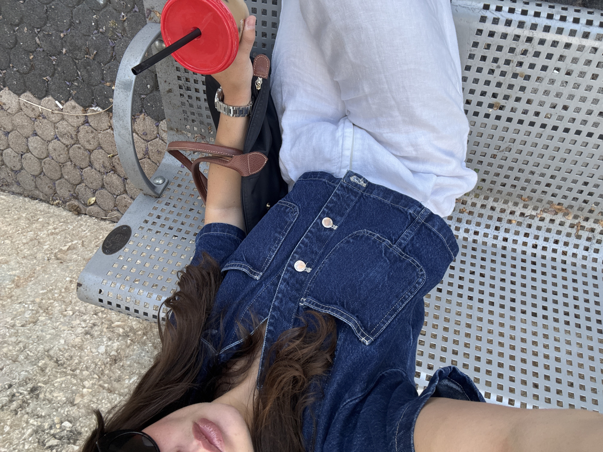 A woman is sitting on a metal bench outdoors, holding a red cup with a straw. She is wearing a denim jacket and white pants. A black bag with brown straps is next to her. The background shows a paved ground with some leaves and dirt. This appears to be an outdoor public space, possibly a park or a street in Tel-Aviv, Israel.