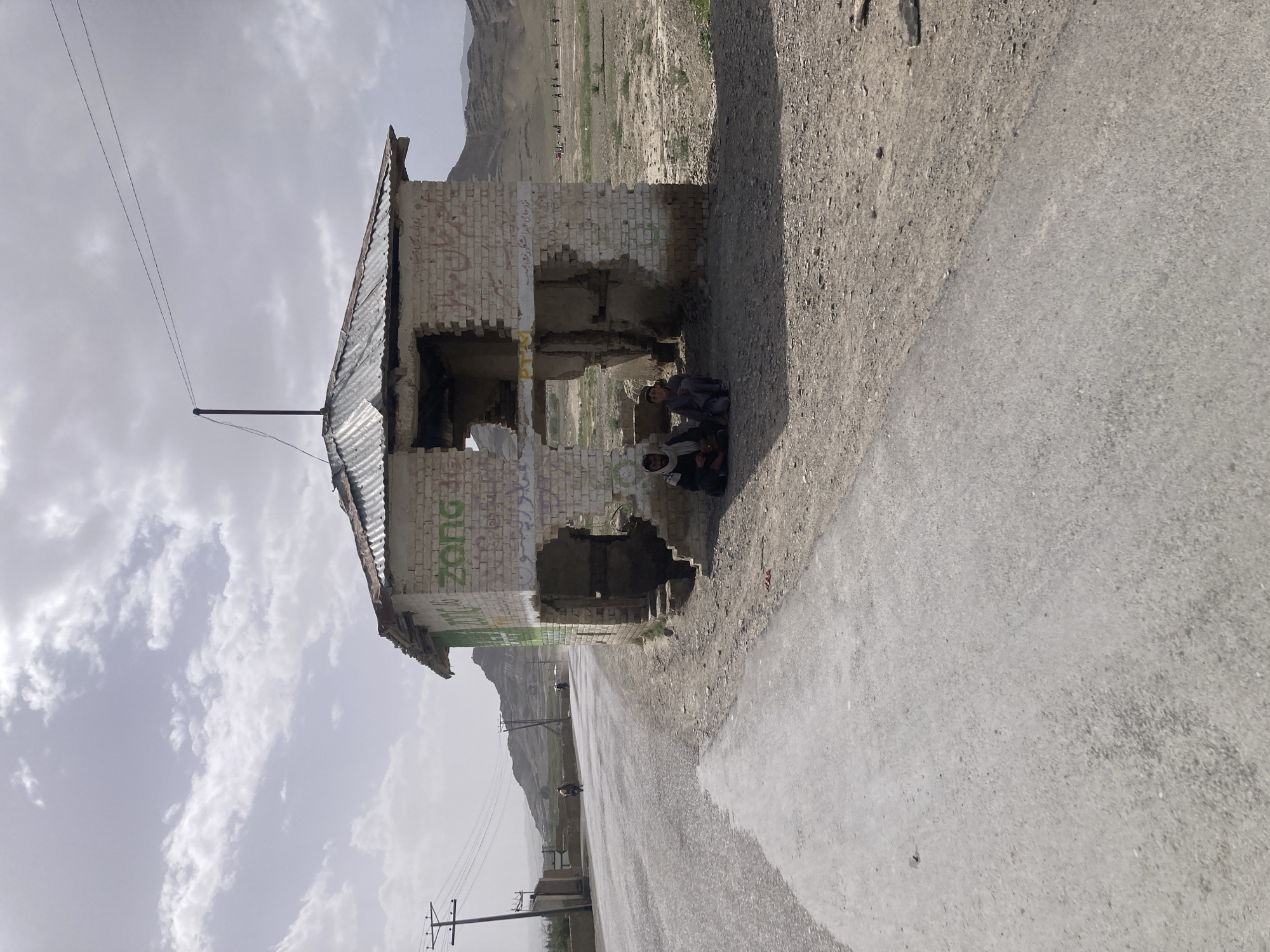 The image shows a dilapidated, graffiti-covered brick structure with a corrugated metal roof, likely a former bus stop, located next to a paved road in what appears to be a rural area. The structure is damaged, with large sections of brick missing. Graffiti, including the word "ZONG" and other unrecognizable markings, are visible on the walls. Two people are sitting inside the structure, seeking shade. The landscape behind the structure is arid, with low hills and sparse vegetation. Electrical poles line the road in the distance. The sky is overcast. The scene is in Gulistan, Pakistan.