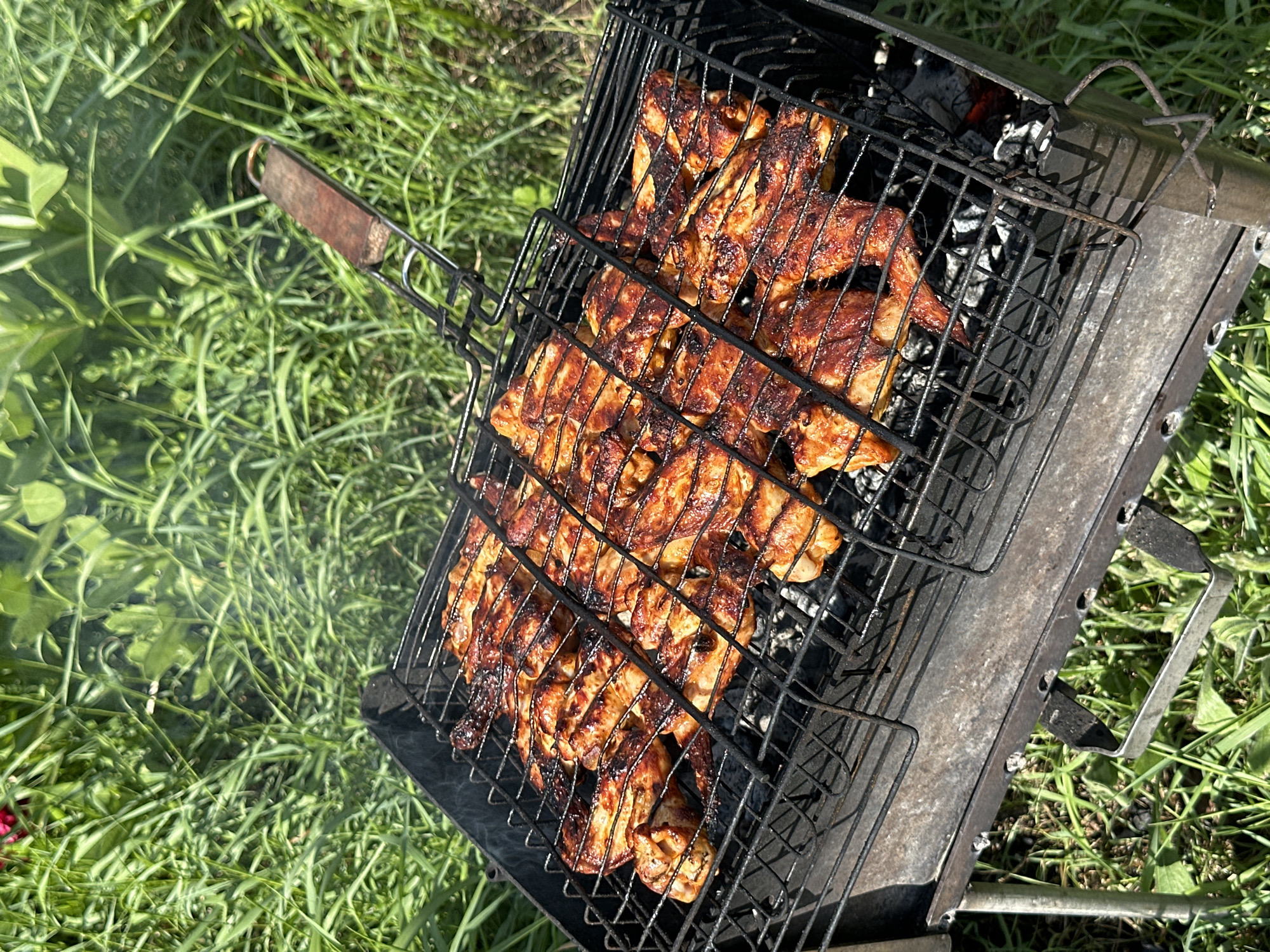 The image depicts a barbecue grill on grass in Khokhryaki, Russia. The grill contains marinated chicken pieces cooking over hot coals. Smoke is visible emanating from the grill. The surrounding environment consists of green grass.