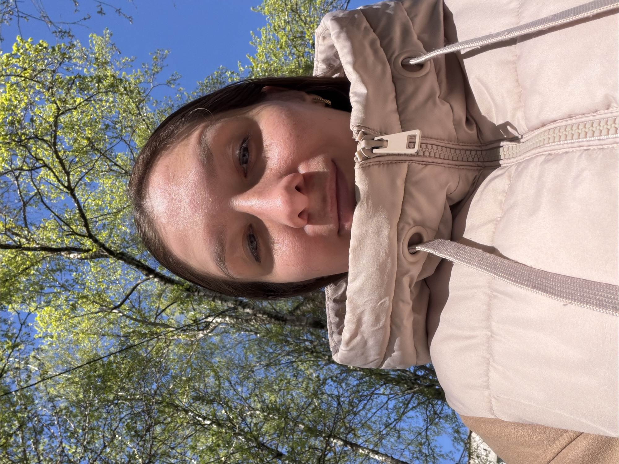 A young woman with light skin is pictured outdoors on a sunny day. She has dark hair and is wearing a light pink hooded jacket. The jacket is zipped up, and the hood is up around her face. The background shows a blue sky with the branches and green leaves of trees filling the upper portion of the image. The photo is taken from a low angle, looking up at the woman. The location is unknown.