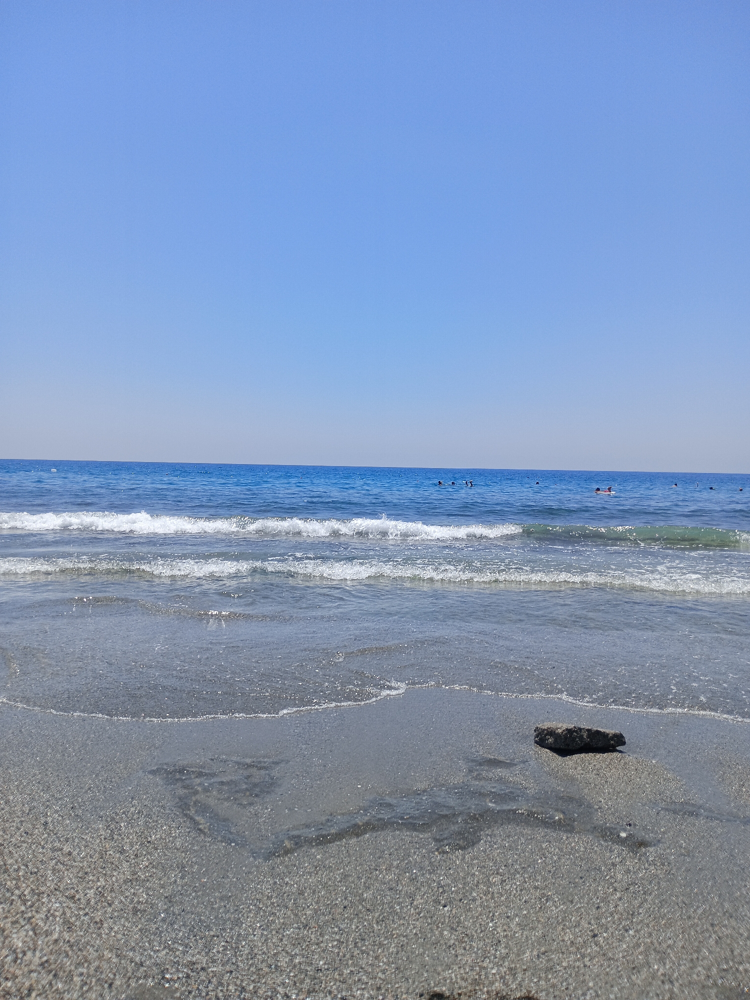 The image depicts a serene beach scene in Alanya, Turkey, under a clear blue sky. The foreground is dominated by wet sand, with gentle waves lapping at the shore. A single, dark-colored rock rests on the wet sand near the center of the frame. In the mid-ground, the clear blue sea stretches towards the horizon, with multiple small, dark figures of people visible in the water, suggesting swimming or other water activities. The horizon line is sharp, separating the calm sea from the expansive sky.