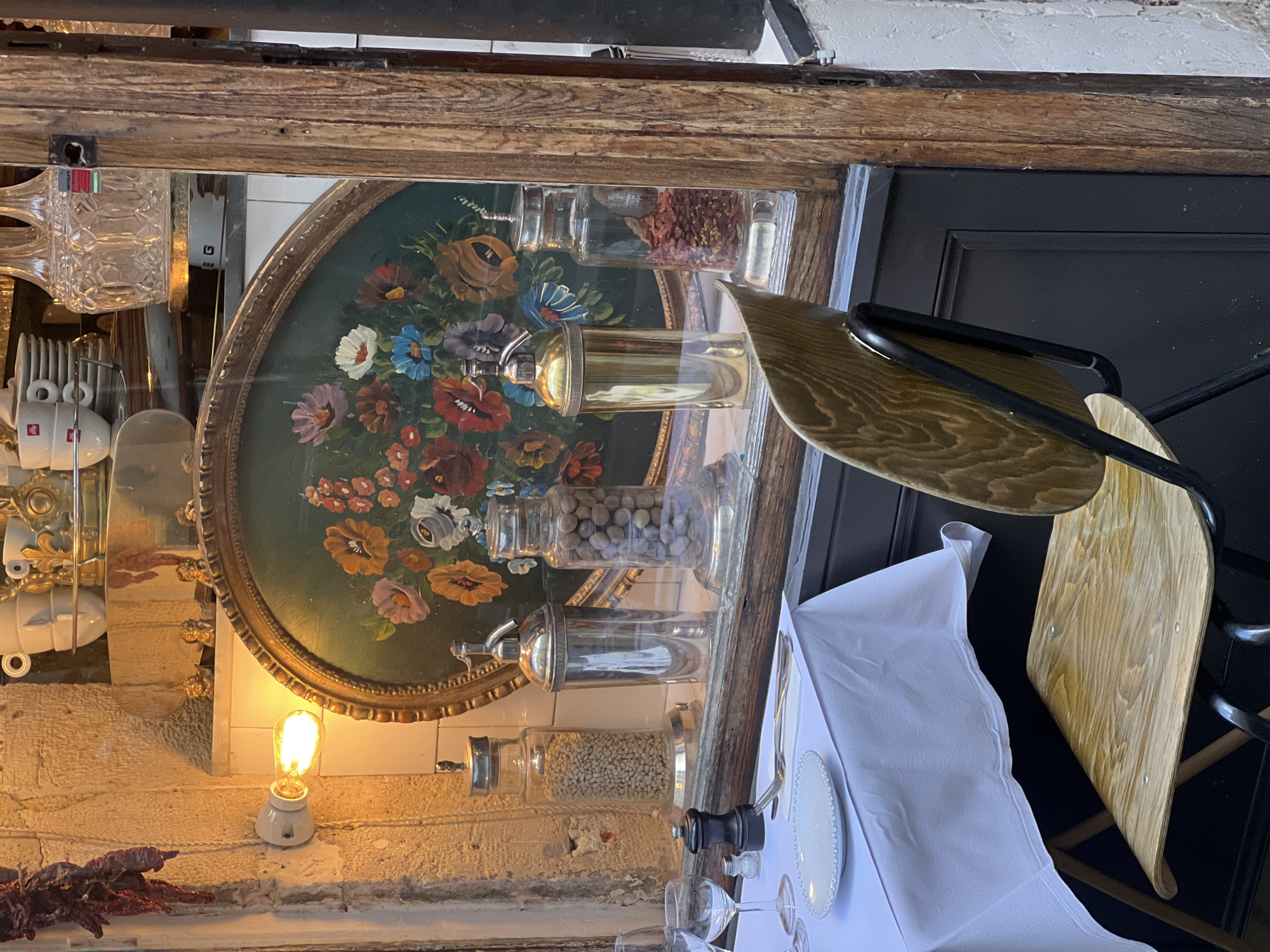 The image depicts a scene inside a restaurant in Paris, France, focusing on details of the interior and table setting.
**Environment:** The interior of the restaurant has a rustic, vintage aesthetic. The walls are textured and painted white or off-white. A wooden beam runs along the ceiling.
**Objects:**
* A large, round, green tray with painted floral designs hangs on the wall, functioning as a decorative element.
* Several glass jars of varying sizes are placed above the tray, containing different food items (nuts, spices, etc.).
* A white table cloth covers the table below, with a simple white plate and silverware on it.
* Several drinking glasses are visible on the table.
* A wooden chair with a black metal frame is positioned near the table.
* A vintage-style light bulb in a white ceramic holder is mounted on the wall.
* Dried red peppers hang near the bottom left corner.
* A crystal pitcher is hung on a wall on the left of the image.
**Subjects:** There are no people visible in the frame.
**Actions:** The scene appears static, depicting a table set for a meal, but currently unoccupied.
**Interactions:** There is no apparent interaction. The objects are arranged in a decorative and functional way.
**Overall:** The image presents a carefully styled scene in a Parisian restaurant, emphasizing vintage details and a refined atmosphere.