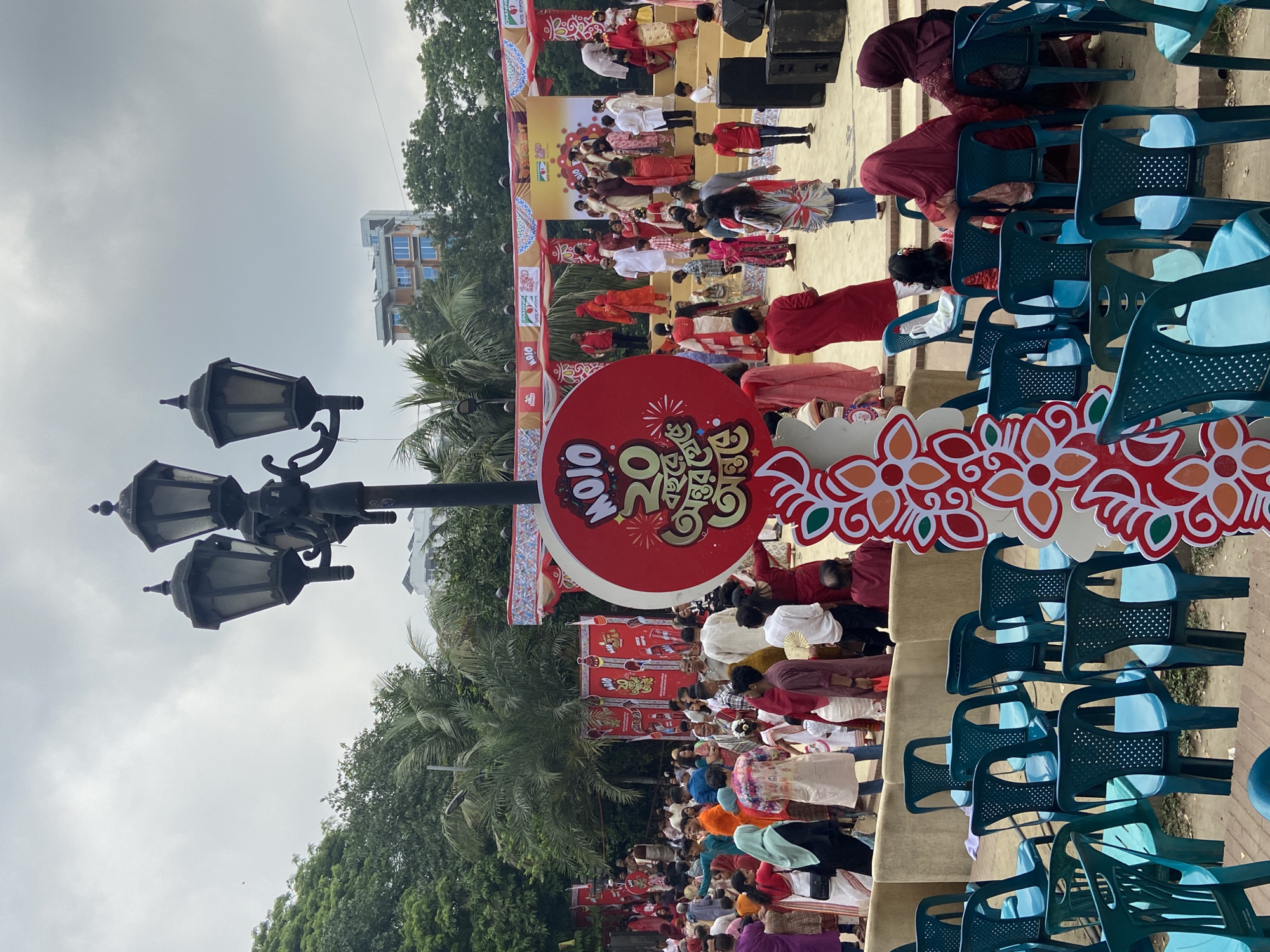 The image depicts a cultural event, likely celebrating Pohela Boishakh, the Bengali New Year, in Dhaka, Bangladesh. A stage is set up with performers in traditional attire, engaging in a musical or dance performance. A crowd is seated in rows of blue plastic chairs, watching the performance. Red and white decorative elements, including a sign with Bengali script, are prominently displayed. A lamppost stands to the side. Palm trees and buildings are visible in the background, suggesting an outdoor location in an urban area. The sky is overcast.