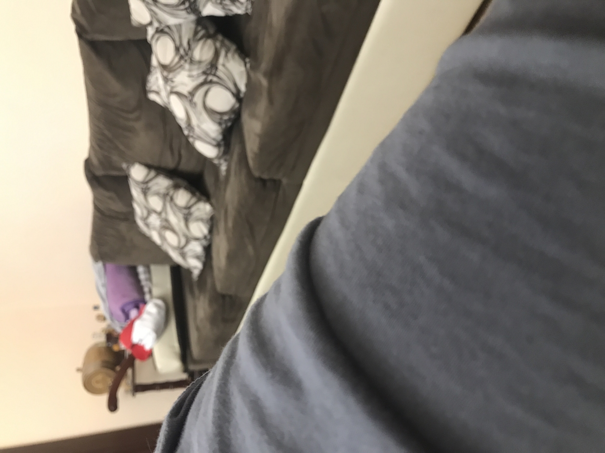 The image shows a living room with a grey couch adorned with black and white patterned pillows. A side table with various items, including a lamp, sits next to the couch. The foreground is dominated by a blurry grey surface, potentially clothing. The location and city of Yerevan, Armenia are not discernible from the image.